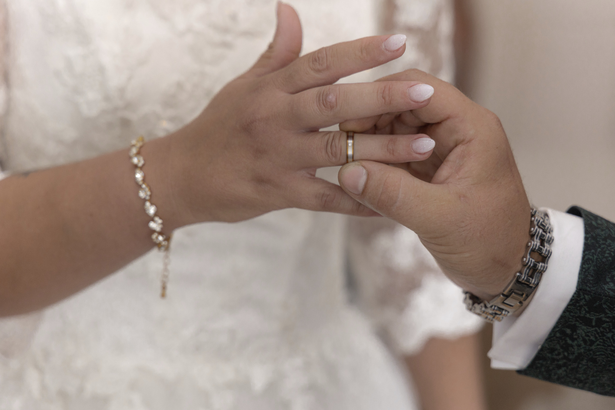 Moment zakładania obrączki ślubnej, dłoń panny młodej z delikatnym manicure i złotą bransoletką, dłoń pana młodego z masywną bransoletą i fragmentem zielonej marynarki.