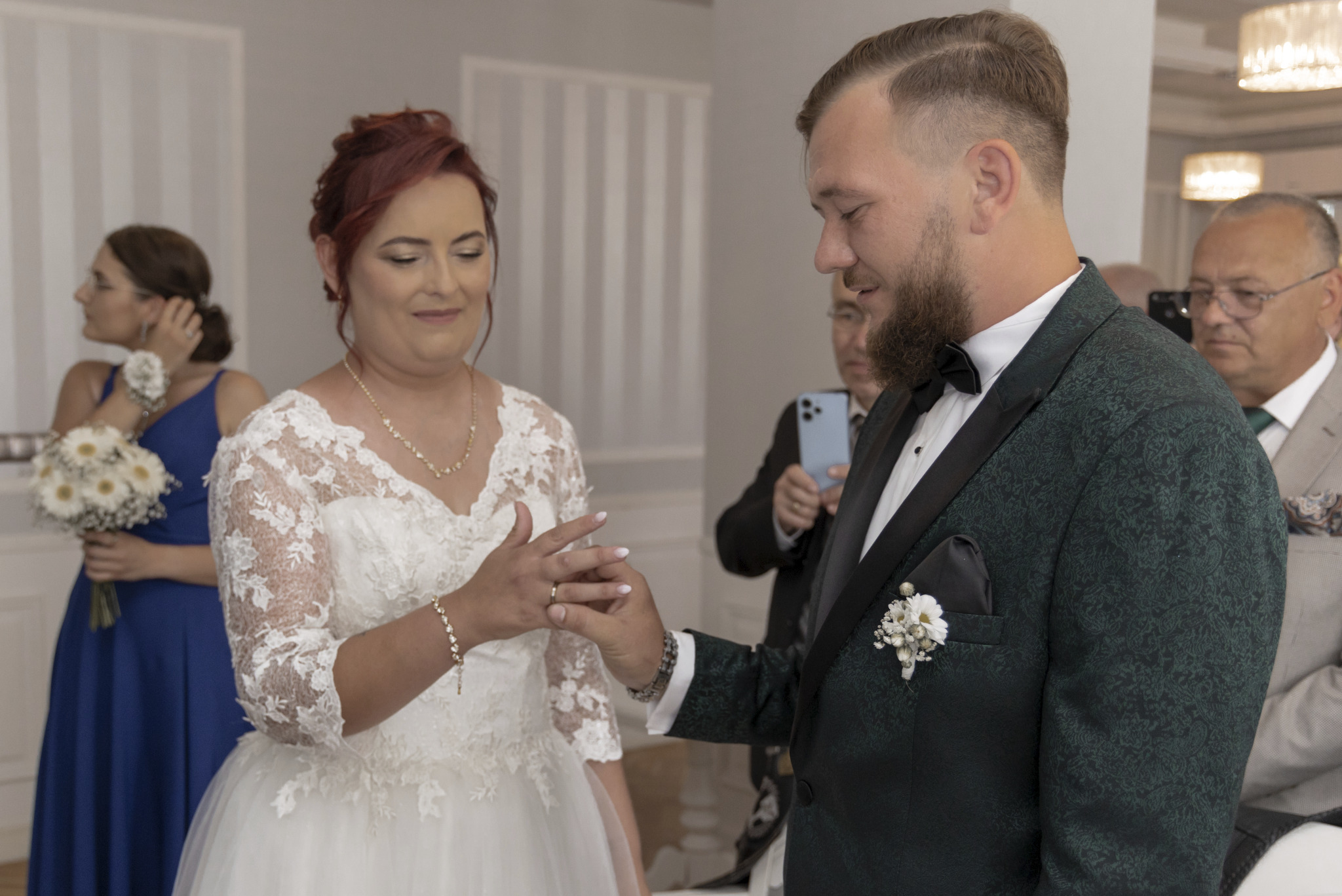 Wymiana obrączek przez parę młodą podczas ceremonii, panna młoda w koronkowej sukni, pan młody w zielonym, wzorzystym smokingu, w tle goście i bukiet kwiatów.