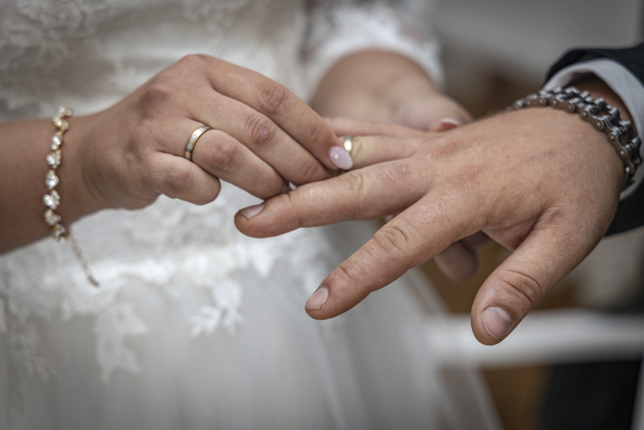 Zbliżenie dłoni z obrączkami podczas ceremonii ślubnej, widoczna delikatna koronka sukni panny młodej i męska bransoletka z ogniw.