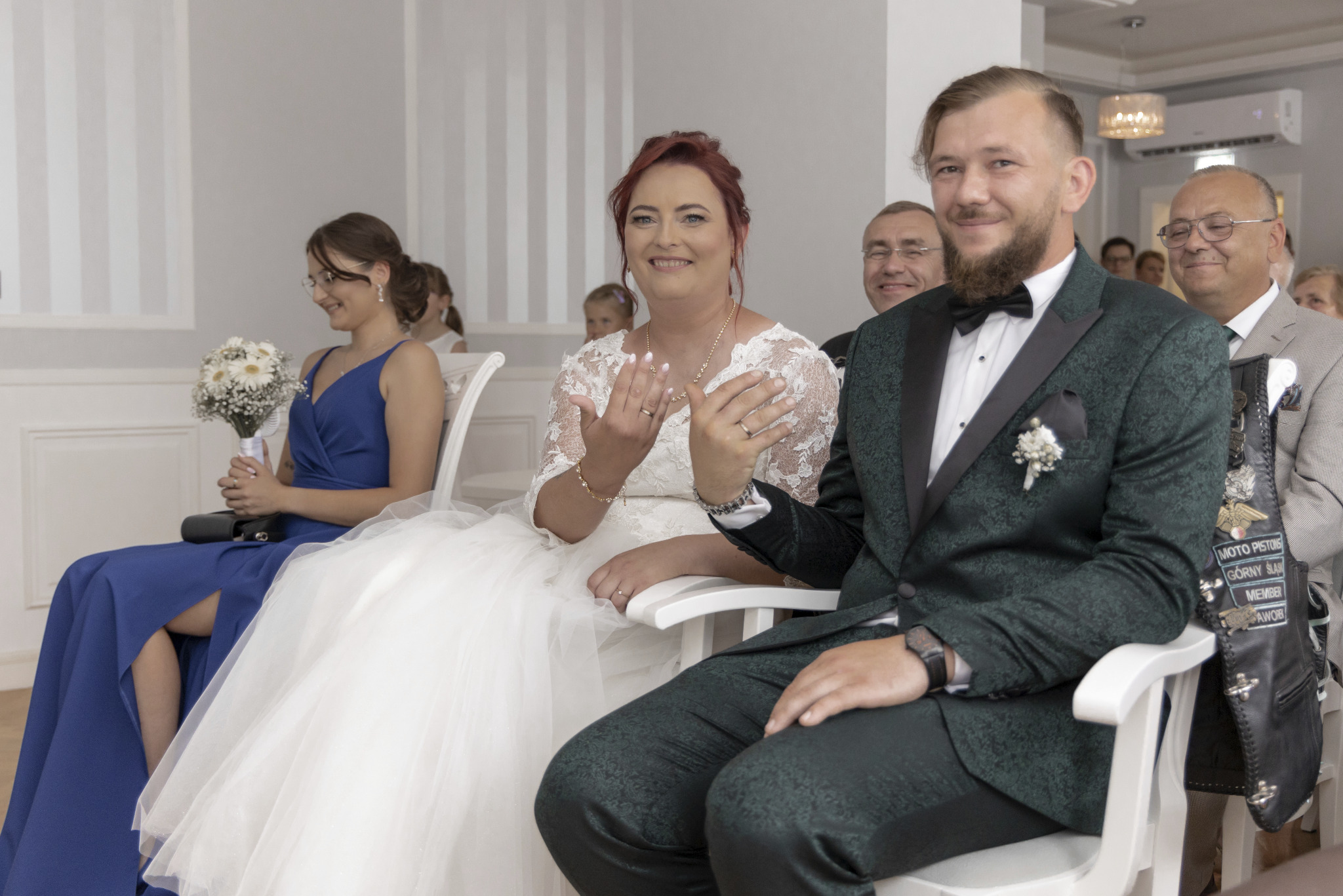 Para młoda siedzi podczas ceremonii ślubnej w Oświęcimiu, panna młoda w koronkowej sukni, pan młody w zielonym, wzorzystym smokingu z muchą i butonierką, goście w tle.