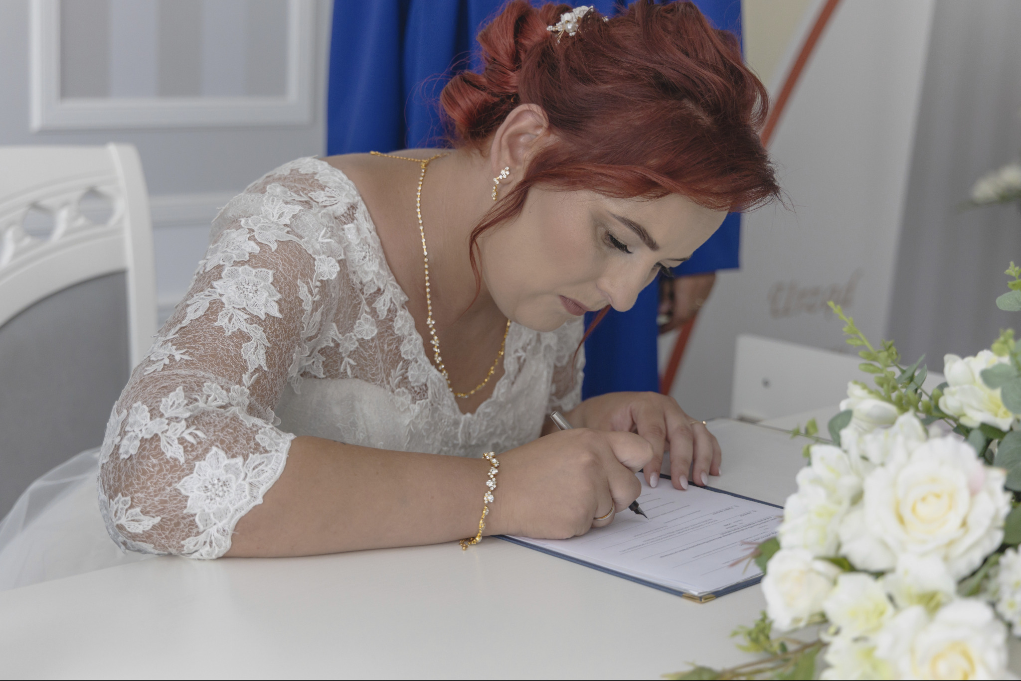 Panna młoda w koronkowej sukni, z upiętymi rudymi włosami i złotą biżuterią, podpisuje dokument na białym stole, obok bukiet białych kwiatów, w tle fragment osoby w niebieskim ubraniu.