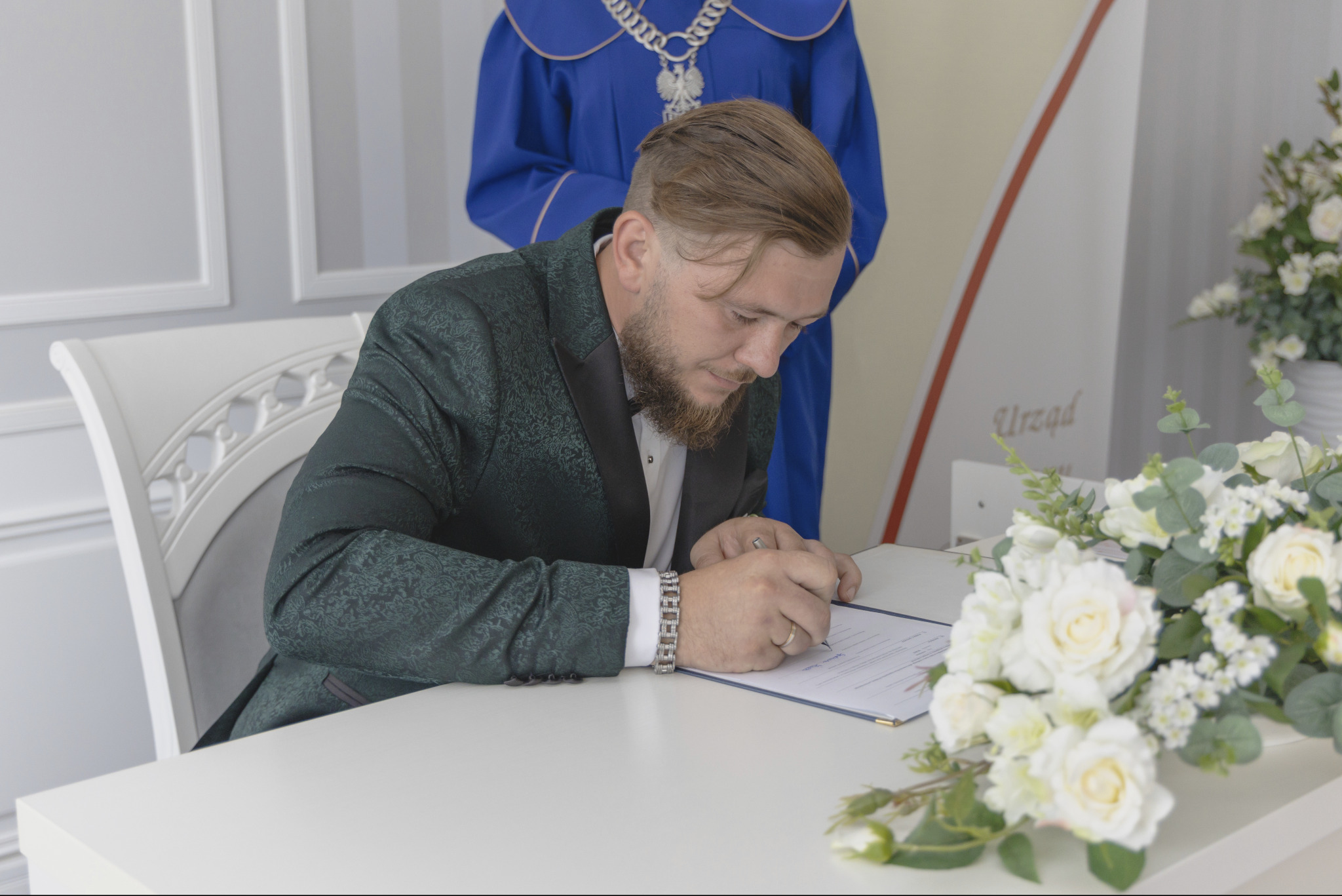 Mężczyzna w zielonym garniturze podpisuje dokument podczas ceremonii ślubnej, widoczny fragment stroju urzędnika i bukiet białych kwiatów.