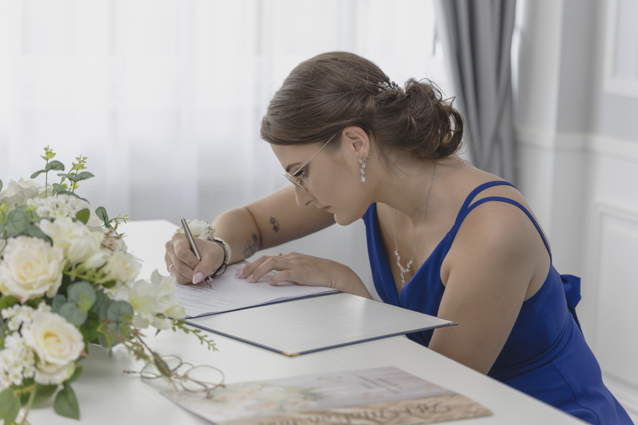Panna młoda w niebieskiej sukience, z bransoletką z kwiatów, podpisuje dokument podczas ceremonii ślubnej, obok bukiet białych róż i odłożone okulary.