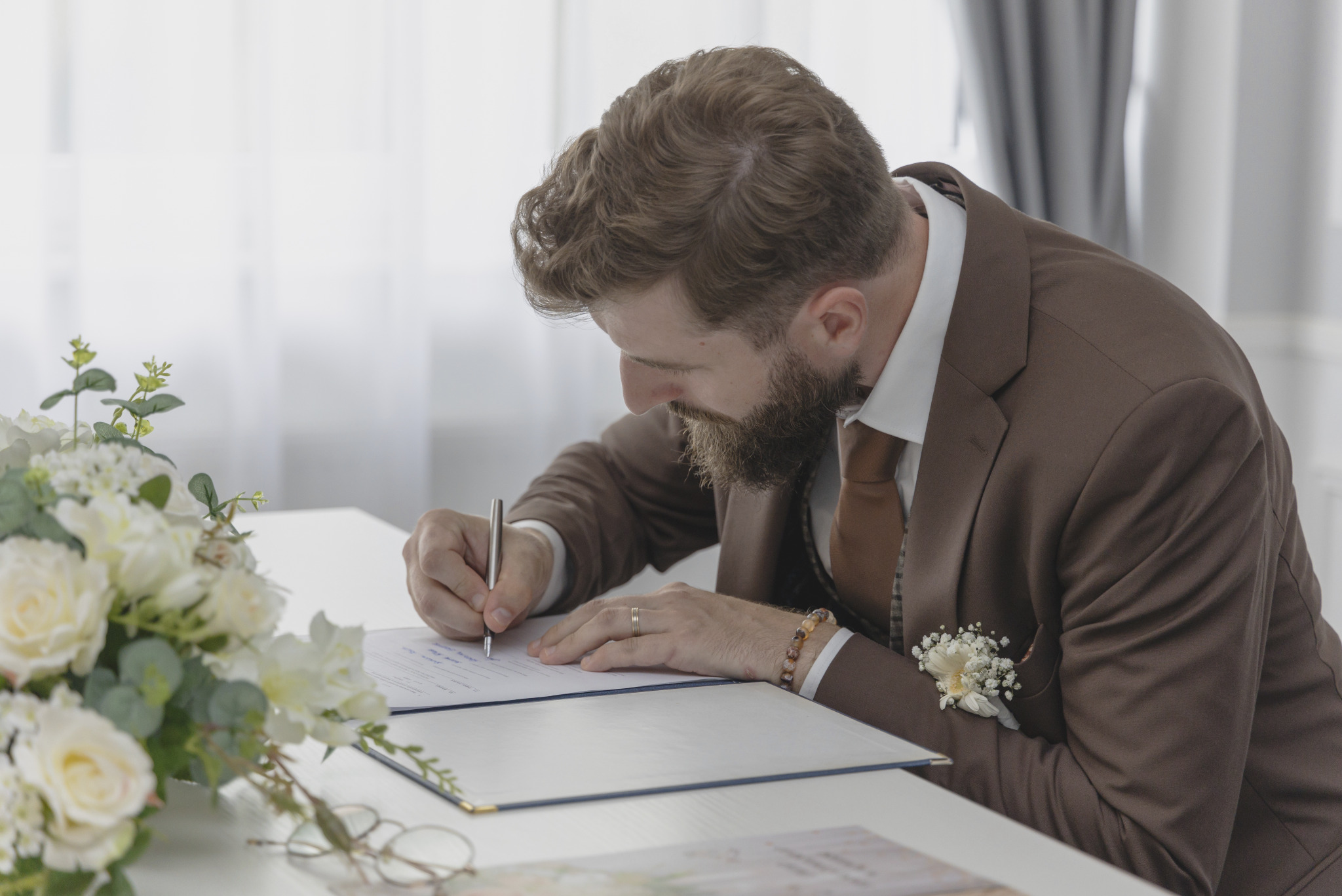 Mężczyzna w brązowym garniturze podpisuje dokument na białym stole z bukietem białych kwiatów. Widoczny pierścionek na palcu i bransoletka z kamieni.