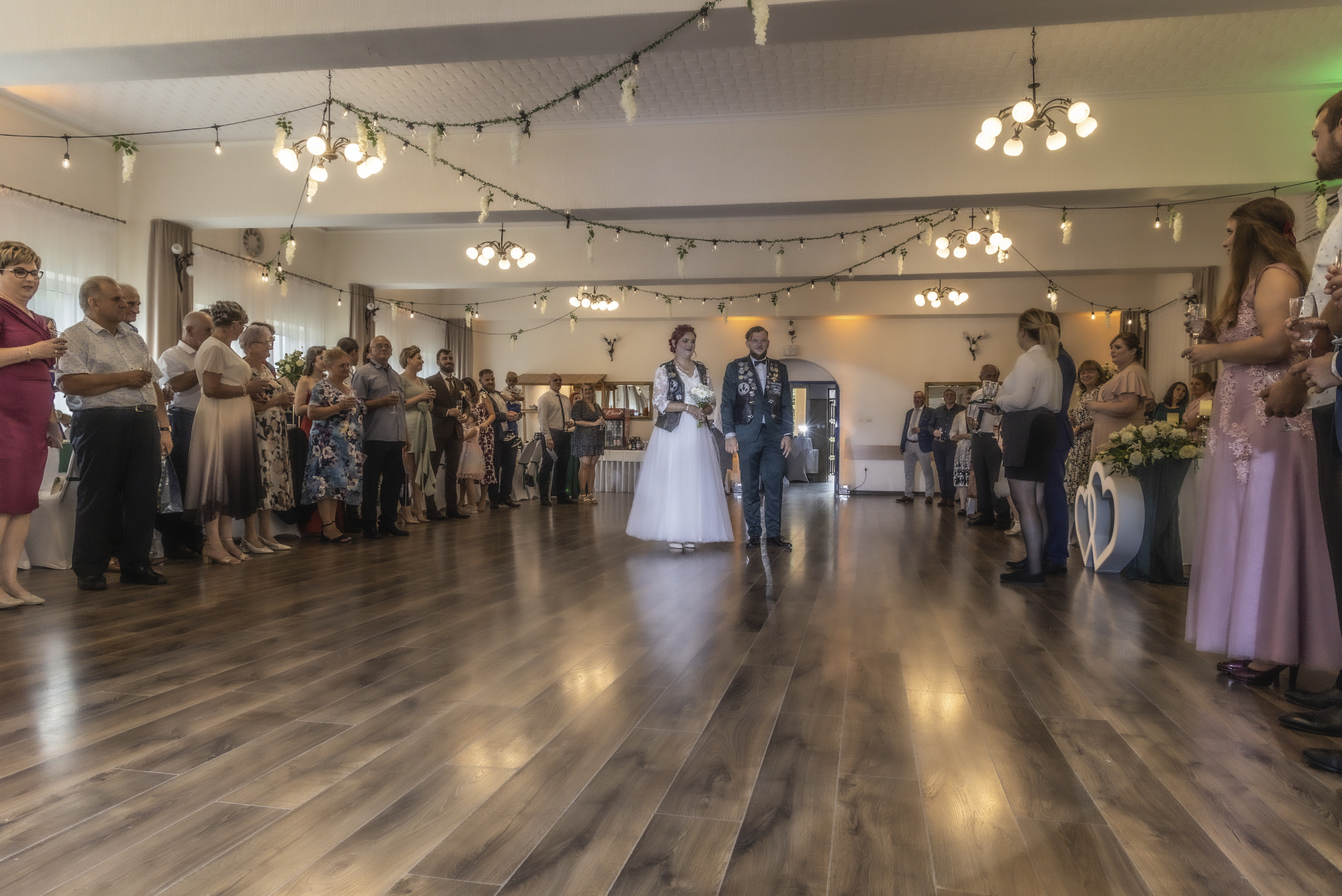 Para młoda w nietypowych strojach ślubnych przechodzi przez szpaler gości w udekorowanej sali z drewnianą podłogą.