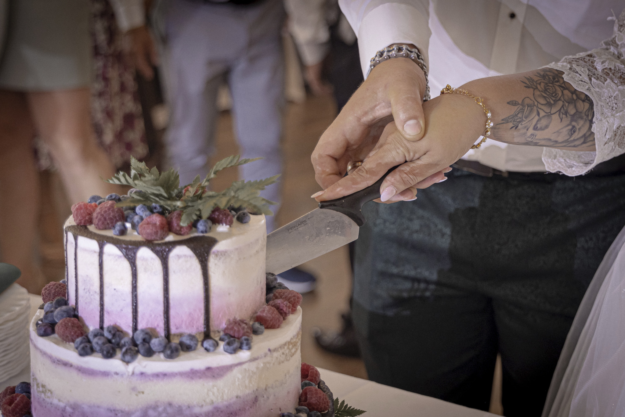 Dwie osoby wspólnie kroją dwupiętrowy tort weselny udekorowany malinami, borówkami i zielonymi listkami; widoczny nóż marki Fiskars.