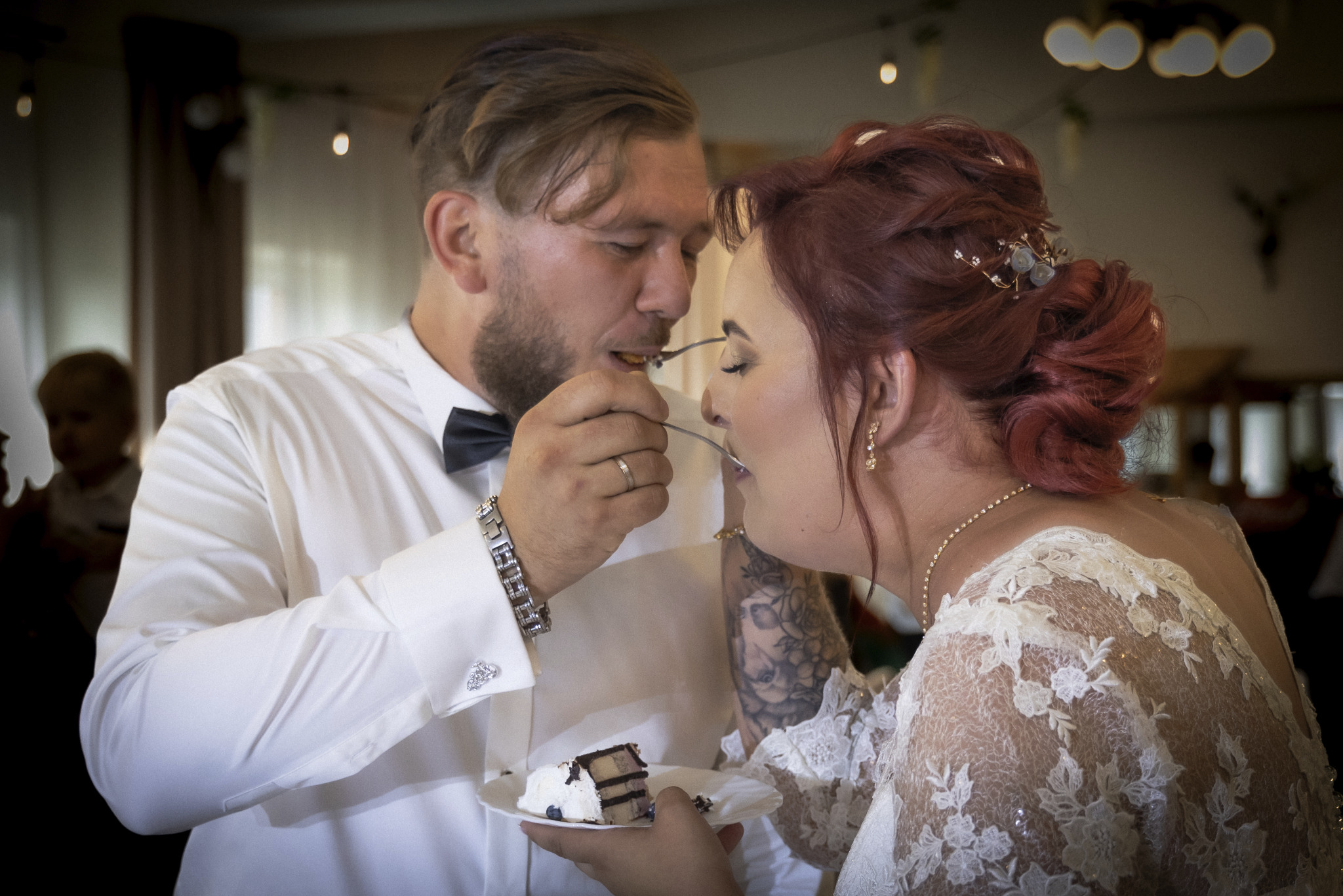 Para młoda w zbliżeniu, karmiąca się nawzajem tortem weselnym. Mężczyzna w białej koszuli z muchą i srebrną bransoletką podaje kawałek tortu kobiecie w koronkowej sukni z upiętymi rudymi włosami.