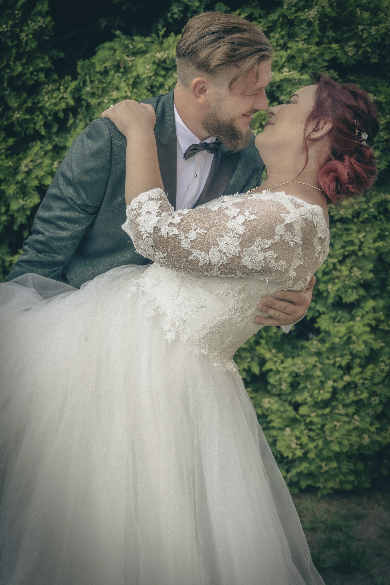Uśmiechnięta para młoda w objęciach na tle zieleni: panna młoda w koronkowej sukni z rękawami 3/4 i upiętymi rudymi włosami, pan młody w szarym smokingu z muszką.