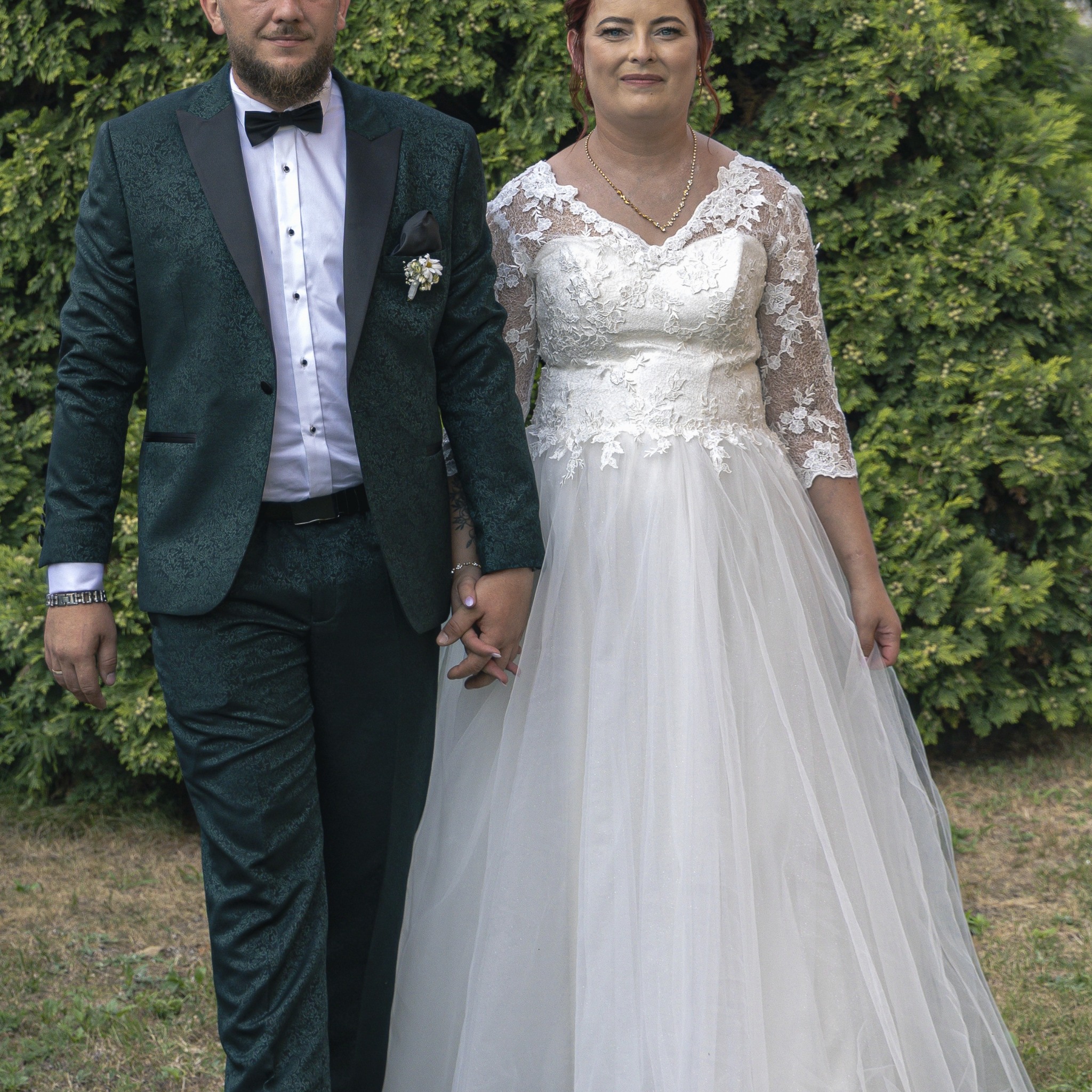 Para młoda trzymająca się za ręce na tle zieleni: pan młody w ciemnozielonym, wzorzystym smokingu z muchą, panna młoda w koronkowej sukni z tiulowym dołem, złoty naszyjnik