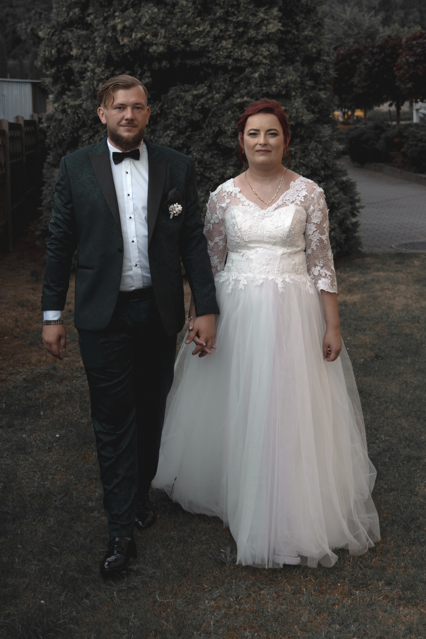 Para młoda, idąca trzymając się za ręce, on w ciemnozielonym garniturze z kwiecistym wzorem, ona w białej sukni ślubnej z koronkową górą i tiulowym dołem na tle zieleni.