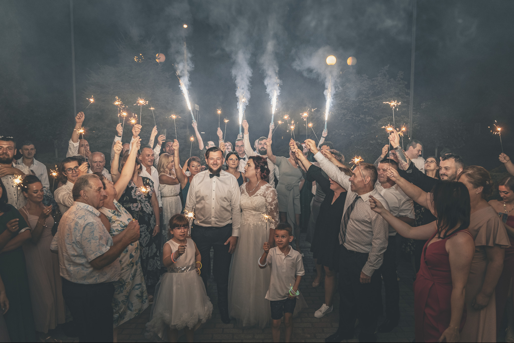 Para młoda otoczona gośćmi weselnymi trzymającymi płonące zimne ognie podczas wieczornego przyjęcia, dzieci w pierwszym rzędzie również z zimnymi ogniami, lekko przyciemnione oświetlenie.