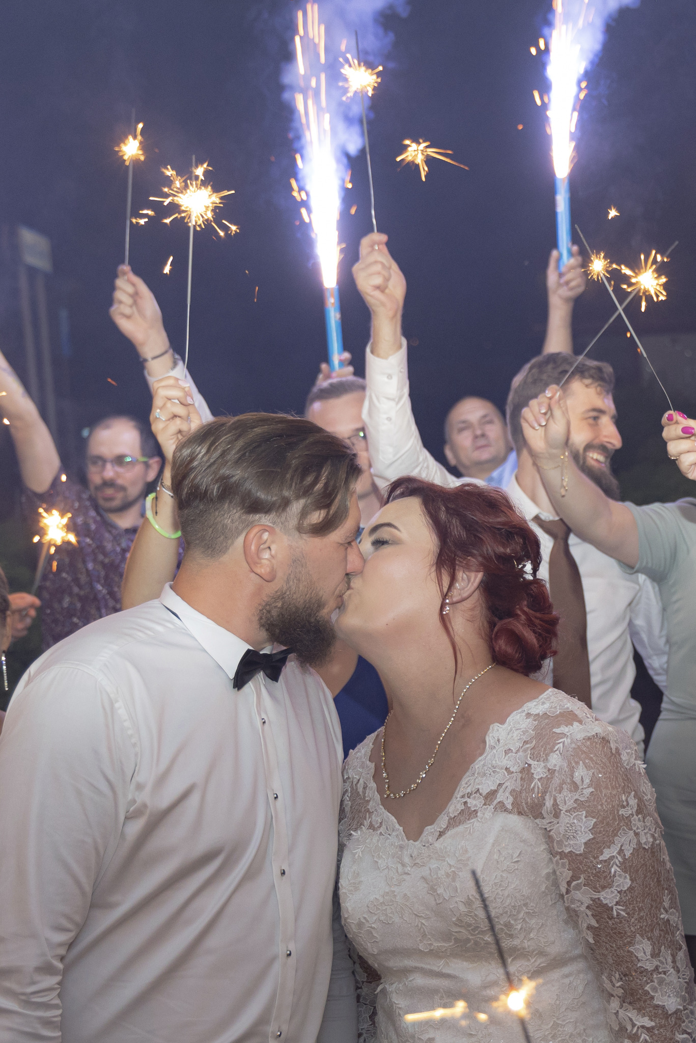 Pocałunek pary młodej otoczonej gośćmi z zimnymi ogniami, panna młoda w koronkowej sukni, pan młody w białej koszuli i muszce, uśmiechnięci goście z iskierkami.