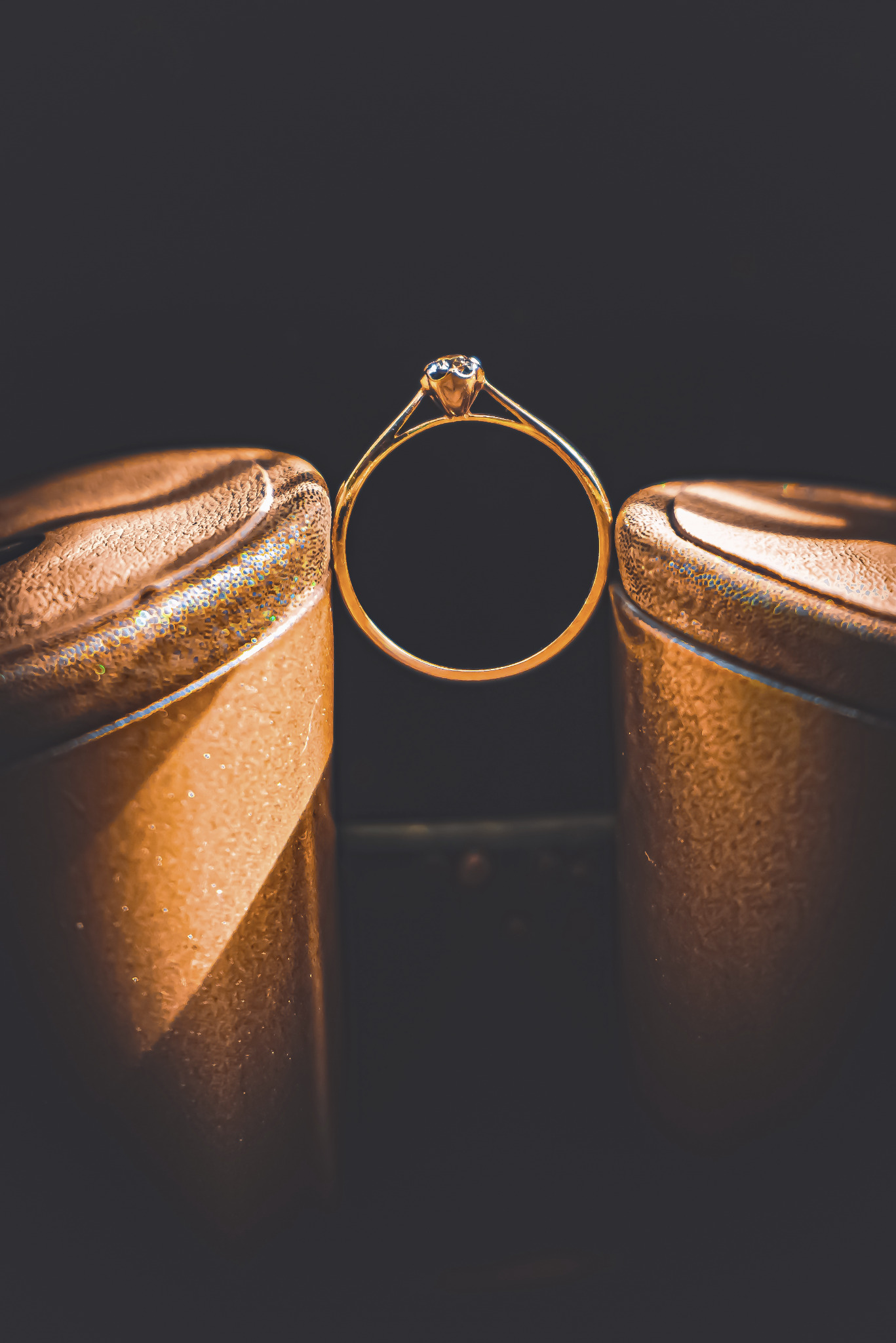 Złoty pierścionek zaręczynowy z brylantem ustawiony między dwoma złotymi szpilkami na ciemnym tle.