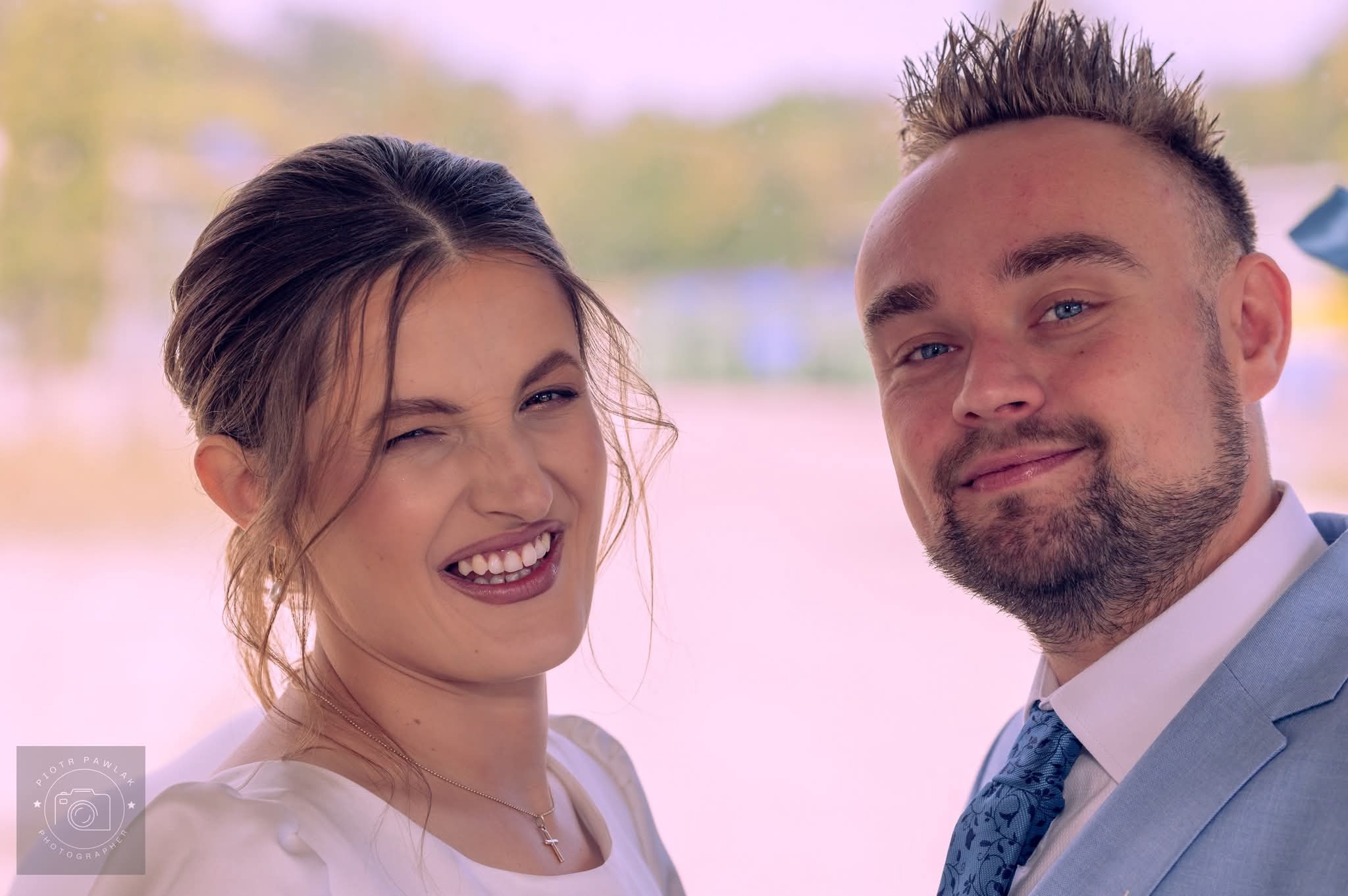 Uśmiechnięta para w zbliżeniu: panna młoda mruga okiem, pan młody w błękitnym garniturze. Widoczny fragment logo fotografa w lewym dolnym rogu.