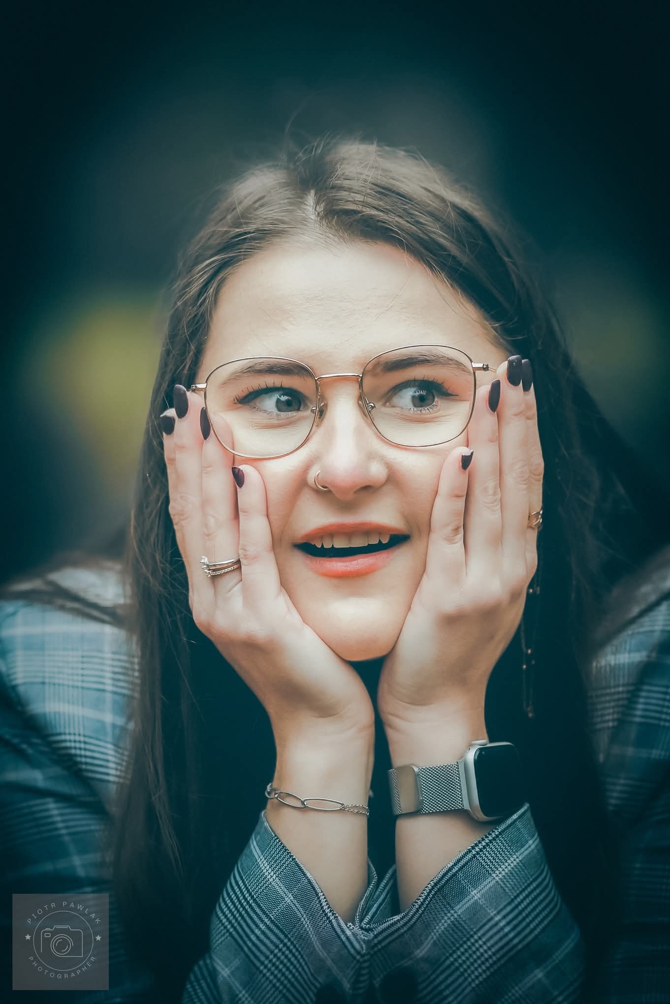 Portret uśmiechniętej kobiety w okularach, z rękami przy twarzy, w bluzie w kratę. Widoczny zegarek na ręce i biżuteria. Ujęcie z bliska, z rozmytym tłem.