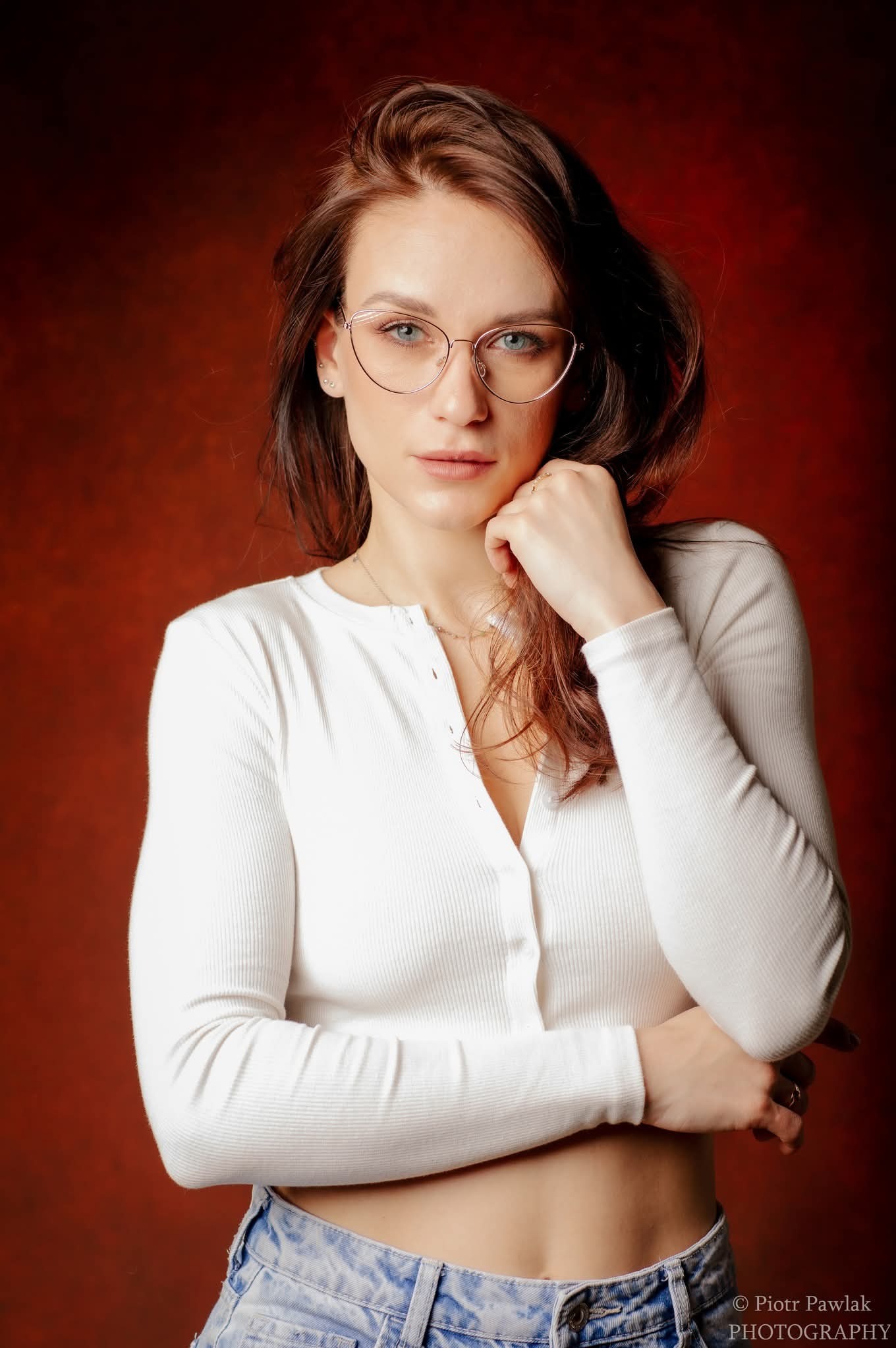 Portret kobiety w okularach, ubrana w białą bluzkę, pozująca na tle czerwonego tła. Delikatny uśmiech, naturalny makijaż, subtelna biżuteria. Ujęcie od pasa w górę.