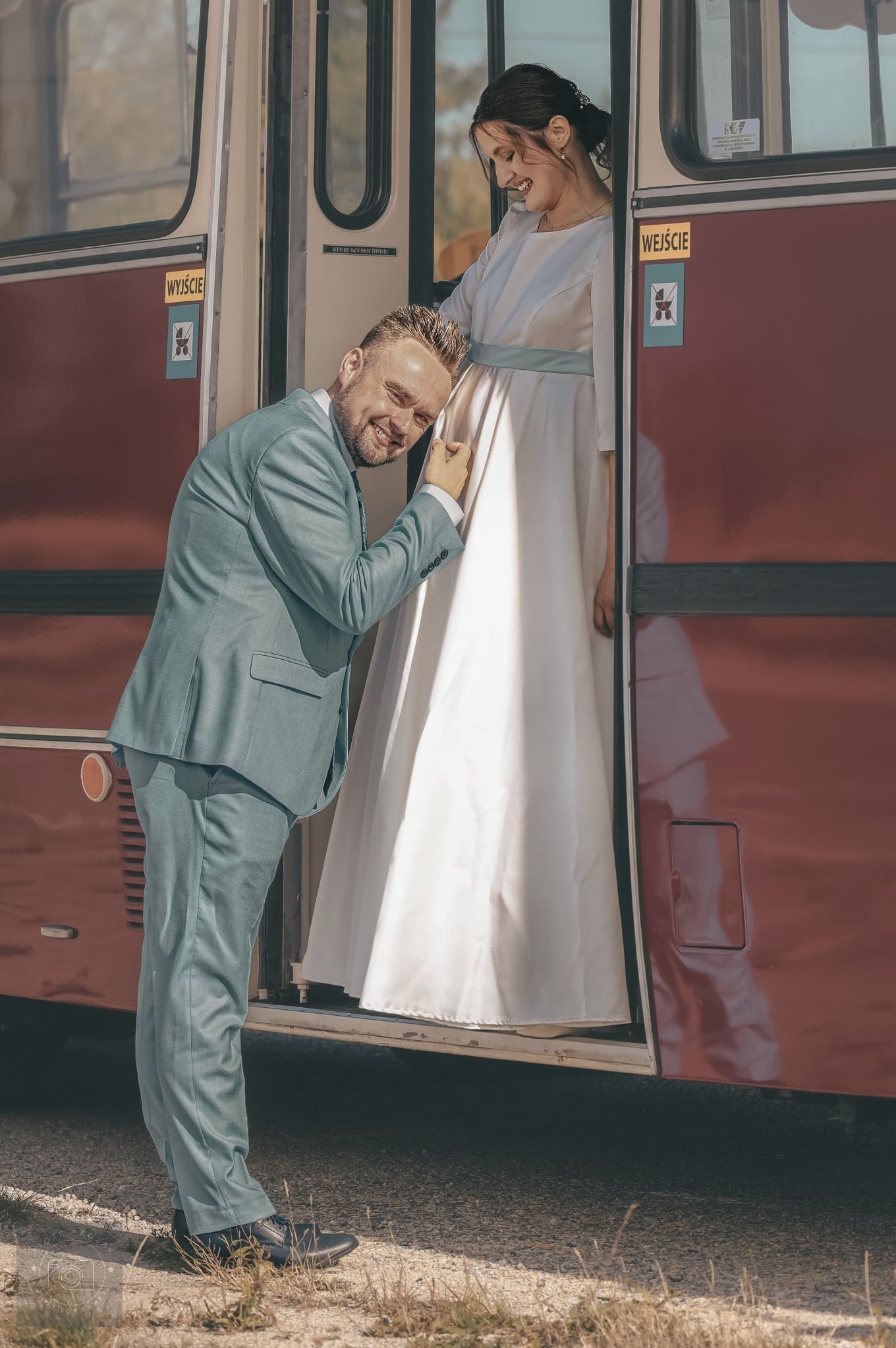 Uśmiechnięty mężczyzna w błękitnym garniturze przykłada ucho do brzucha kobiety w jasnej sukni, stojącej w drzwiach czerwonego tramwaju. Nietypowa sesja ciążowa.