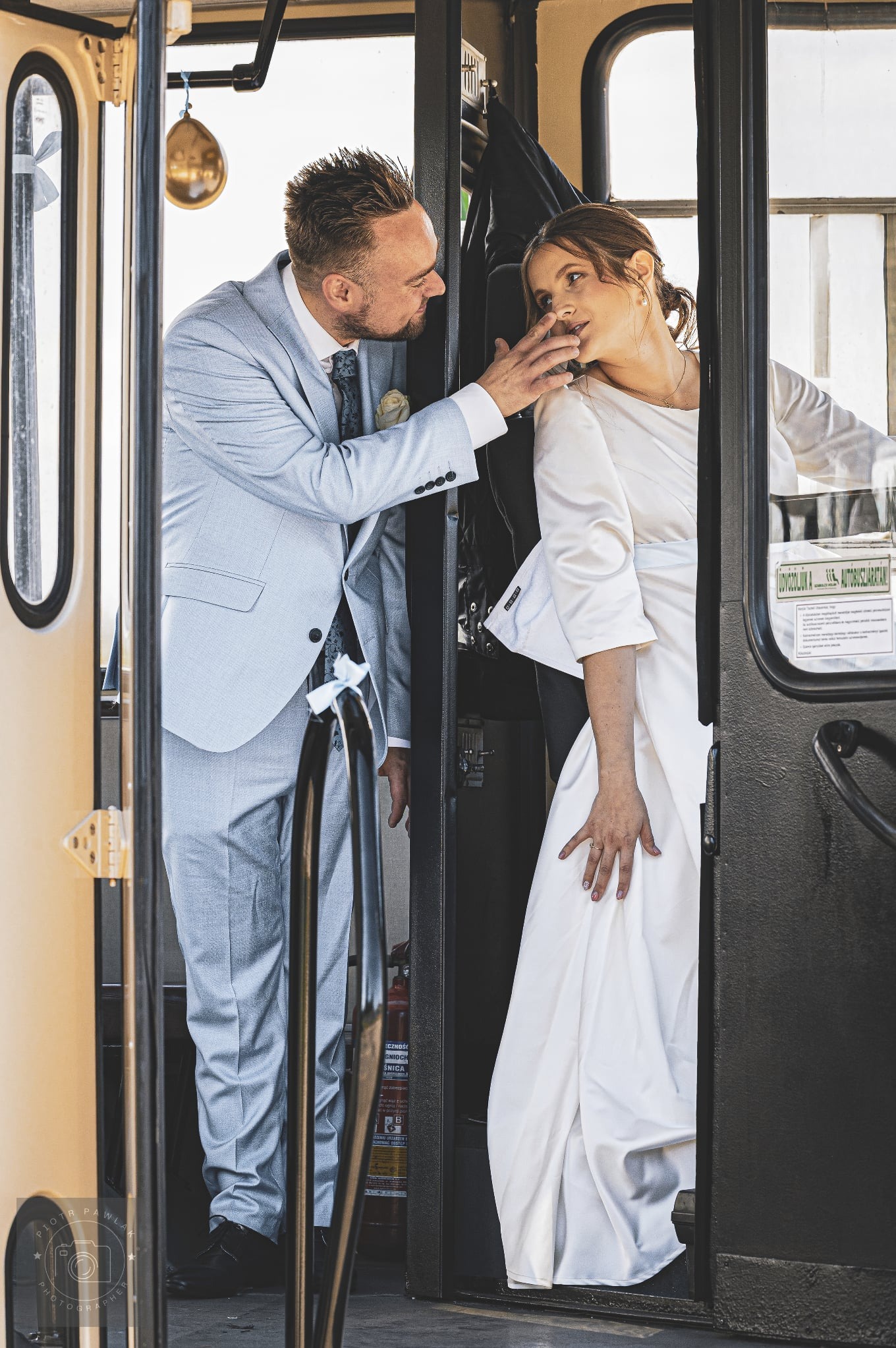 Para młoda w tramwaju: pan młody w błękitnym garniturze dotyka twarzy panny młodej w białej sukni. Nietypowa sceneria ślubna z gaśnicą w tle.