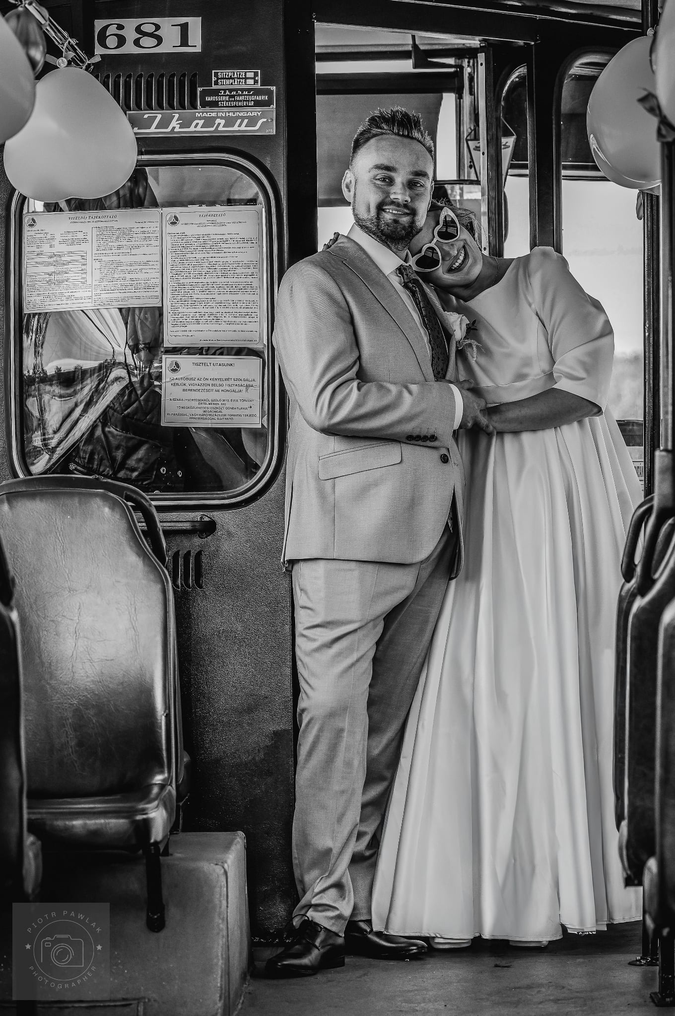 Czarno-białe zdjęcie uśmiechniętej pary młodej w tramwaju Ikarus. Panna młoda w okularach w kształcie serc opiera głowę na ramieniu pana młodego.