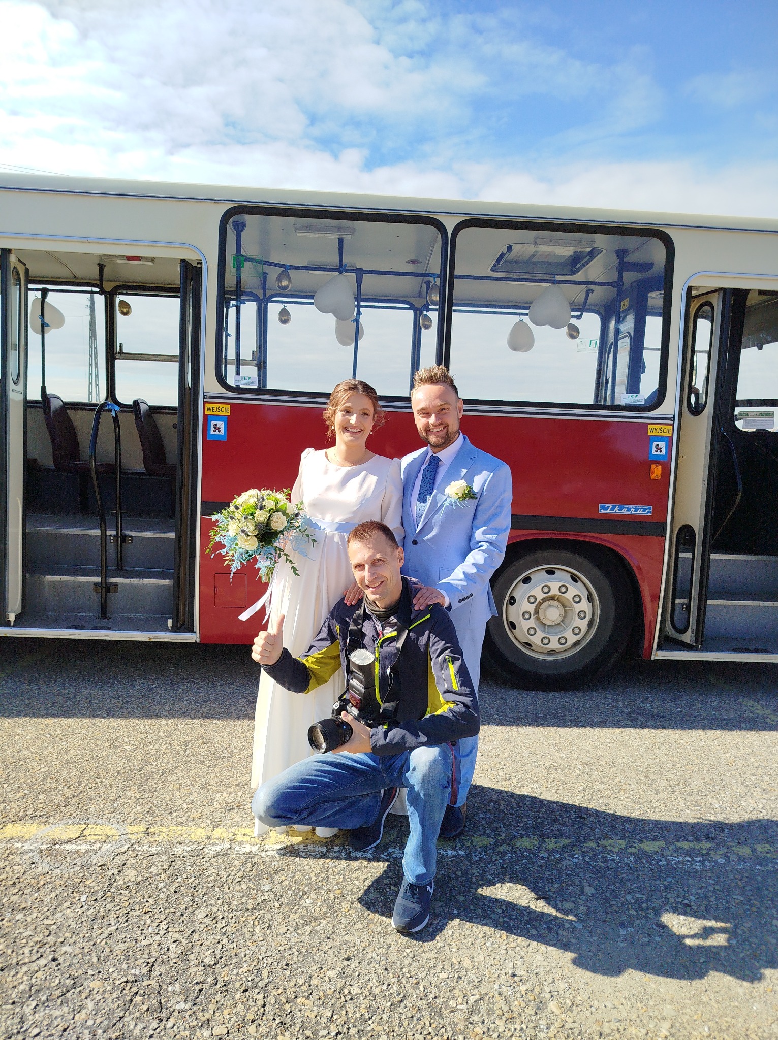 Uśmiechnięty fotograf z parą młodą pozuje na tle czerwonego autobusu Ikarus. Fotograf kuca, trzyma aparat i pokazuje kciuk w górę. Słoneczny dzień.