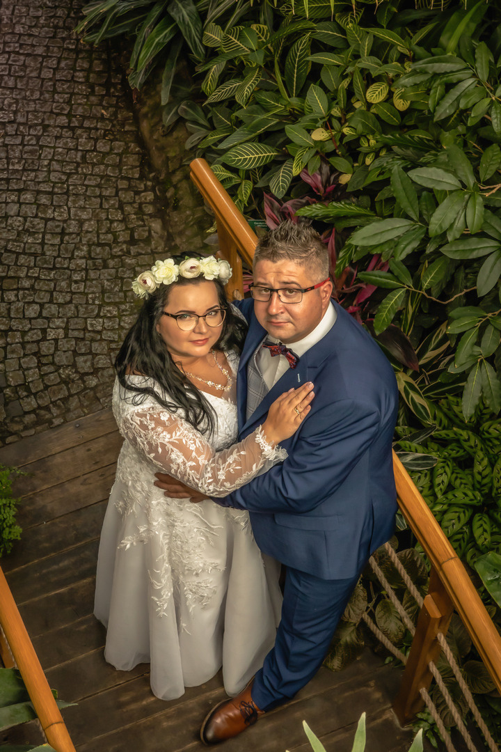 Para młoda w objęciach, ona w koronie z białych kwiatów i sukni z koronkowymi rękawami, on w granatowym garniturze i bordowej muszce, na tle egzotycznej roślinności i kamiennej ścieżki, ujęcie z góry.