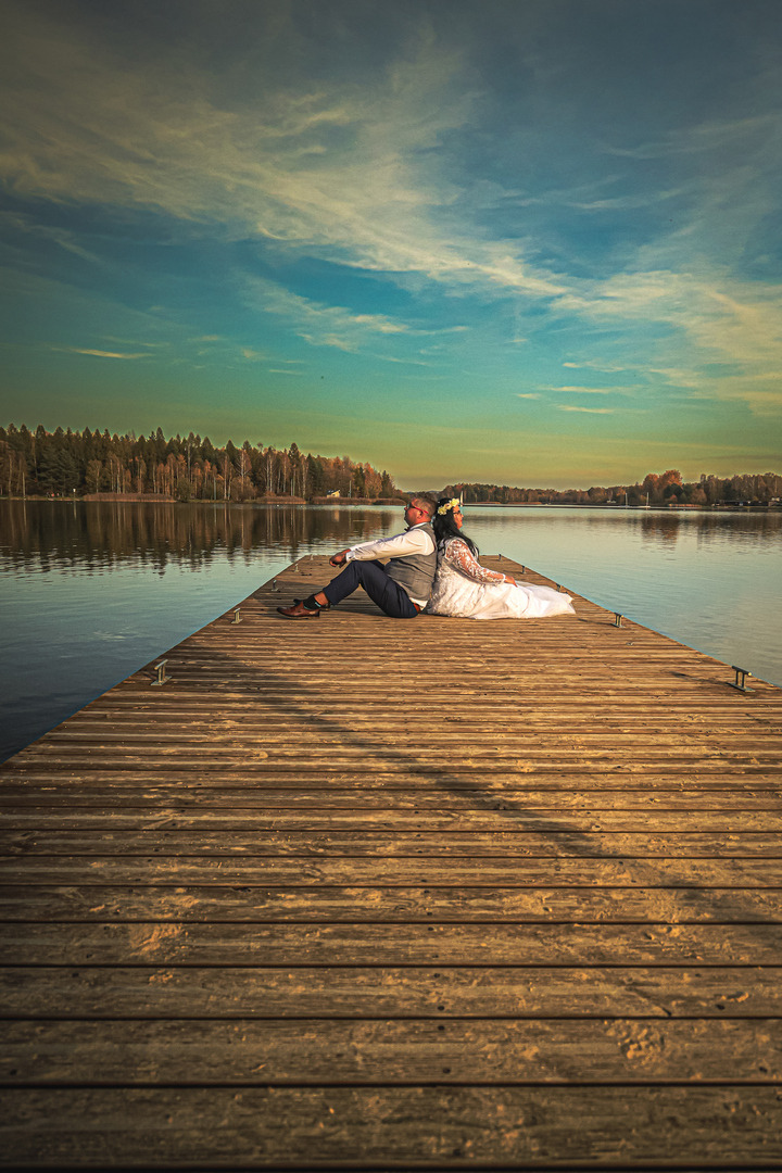 Para siedzi plecami do siebie na drewnianym molo nad jeziorem, w tle las i niebo z delikatnymi chmurami, panna młoda ma wianek na głowie.