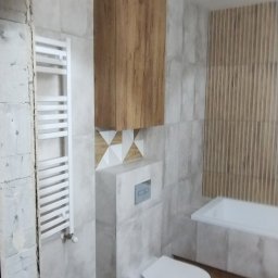 Euro styl wykończenia wnętrz Stanisław Moskwa - Wykończona łazienka z wiszącą szafką z drewnianym frontem, grzejnikiem drabinkowym, toaletą i wanną, ściany wyłożone płytkami imitującymi kamień oraz dekoracyjnymi lamelami.