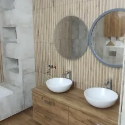 Nowoczesna łazienka z dwiema okrągłymi umywalkami nablatowymi, drewnianą szafką pod umywalki z dwoma szufladami oraz dwoma okrągłymi lustrami w szarej ramie na ścianie z drewnopodobnych paneli...