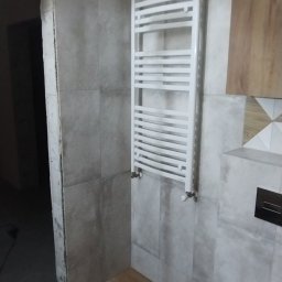 Euro styl wykończenia wnętrz Stanisław Moskwa - Łazienka w trakcie remontu: szare płytki, biały grzejnik drabinkowy, widoczne niedokończone wykończenia przy ościeżnicy.