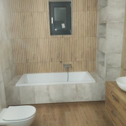 Euro styl wykończenia wnętrz Stanisław Moskwa - Nowoczesna łazienka z drewnopodobnymi płytkami na ścianie i podłodze, biała wanna pod oknem, wisząca toaleta i umywalka na drewnianej szafce.