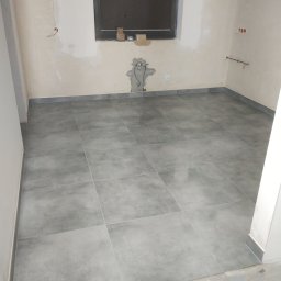 Euro styl wykończenia wnętrz Stanisław Moskwa - Szare płytki podłogowe ułożone w kwadraty w pustym pomieszczeniu z oknem i instalacjami na ścianach, widoczne niedokończone wykończenie ścian i podłogi.