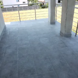 Szare płytki gresowe ułożone na balkonie z metalową balustradą i widokiem na domy w oddali.