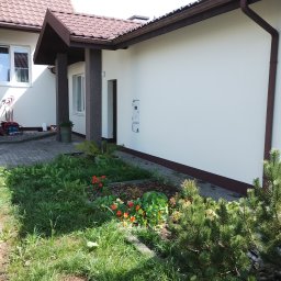 Euro styl wykończenia wnętrz Stanisław Moskwa - Jasna elewacja domu jednorodzinnego z brązowym dachem i elementami wykończeniowymi, widok z poziomu ogrodu z kwitnącymi nasturcjami.