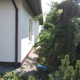 Biała elewacja domu jednorodzinnego w Strzyżowie z brązowymi akcentami, widoczna z poziomu ogrodu. Ścieżka z kostki prowadzi wzdłuż ściany.