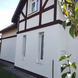 Odnowiona elewacja domu w stylu szachulcowym, z kremową ścianą i brązowymi belkami. Widoczne okna i fragment chodnika. Całość w słoneczny dzień.