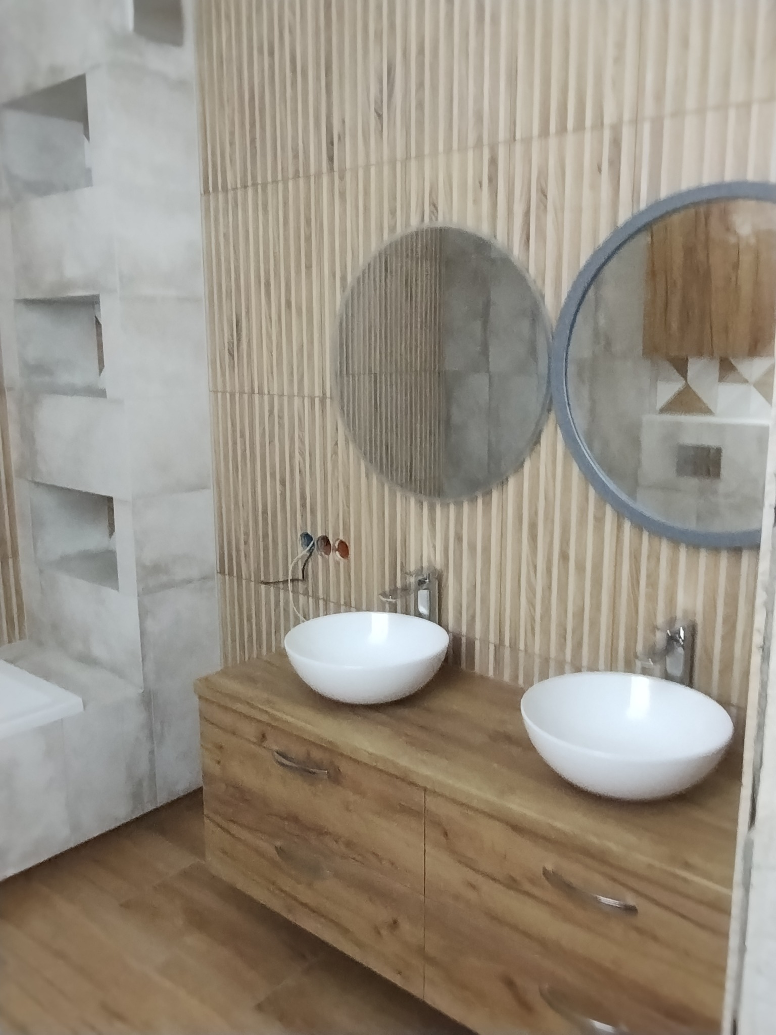 Nowoczesna łazienka z dwiema okrągłymi umywalkami nablatowymi, drewnianą szafką pod umywalki z dwoma szufladami oraz dwoma okrągłymi lustrami w szarej ramie na ścianie z drewnopodobnych paneli...