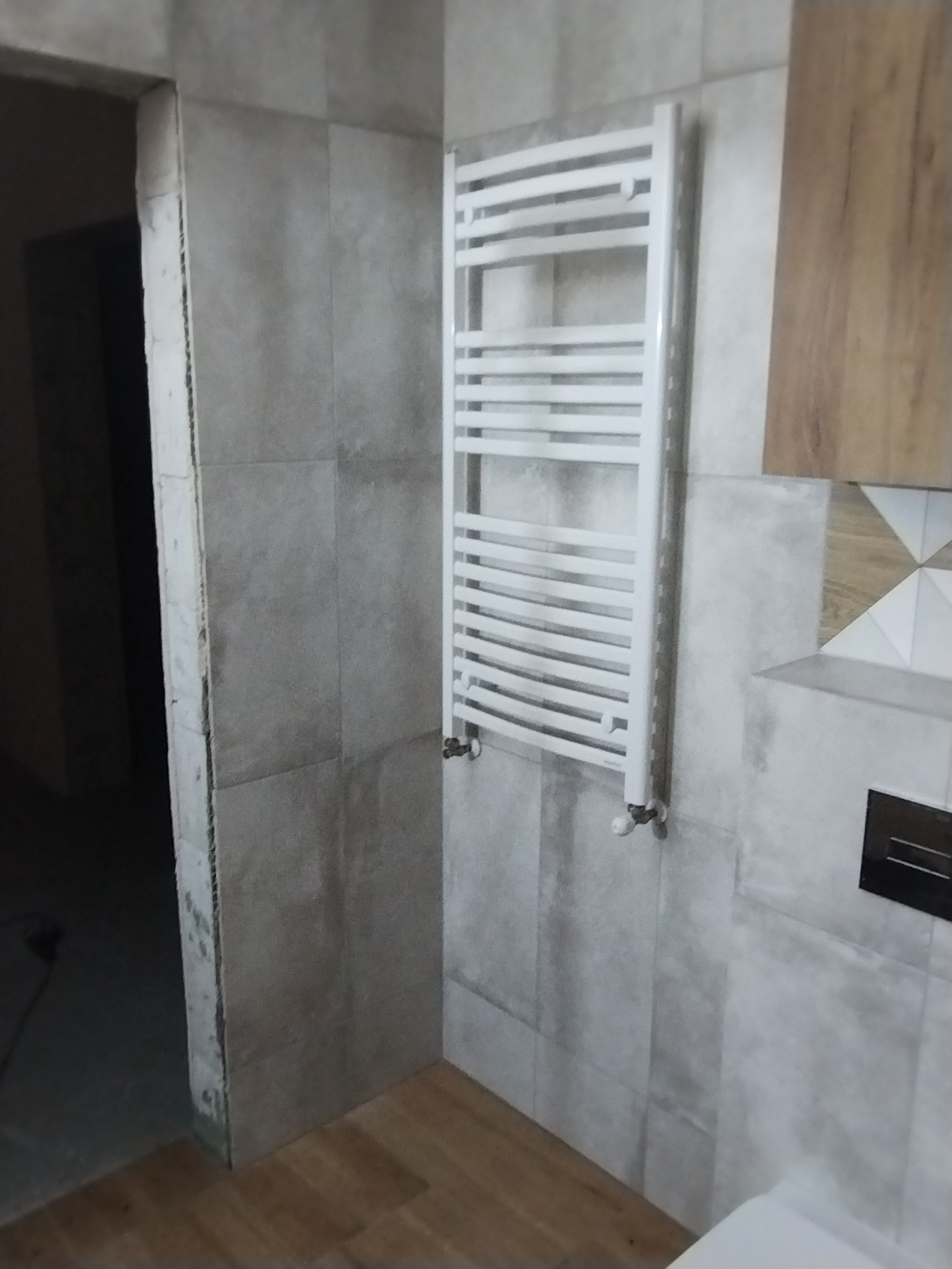 Łazienka w trakcie remontu: szare płytki, biały grzejnik drabinkowy, widoczne niedokończone wykończenia przy ościeżnicy.