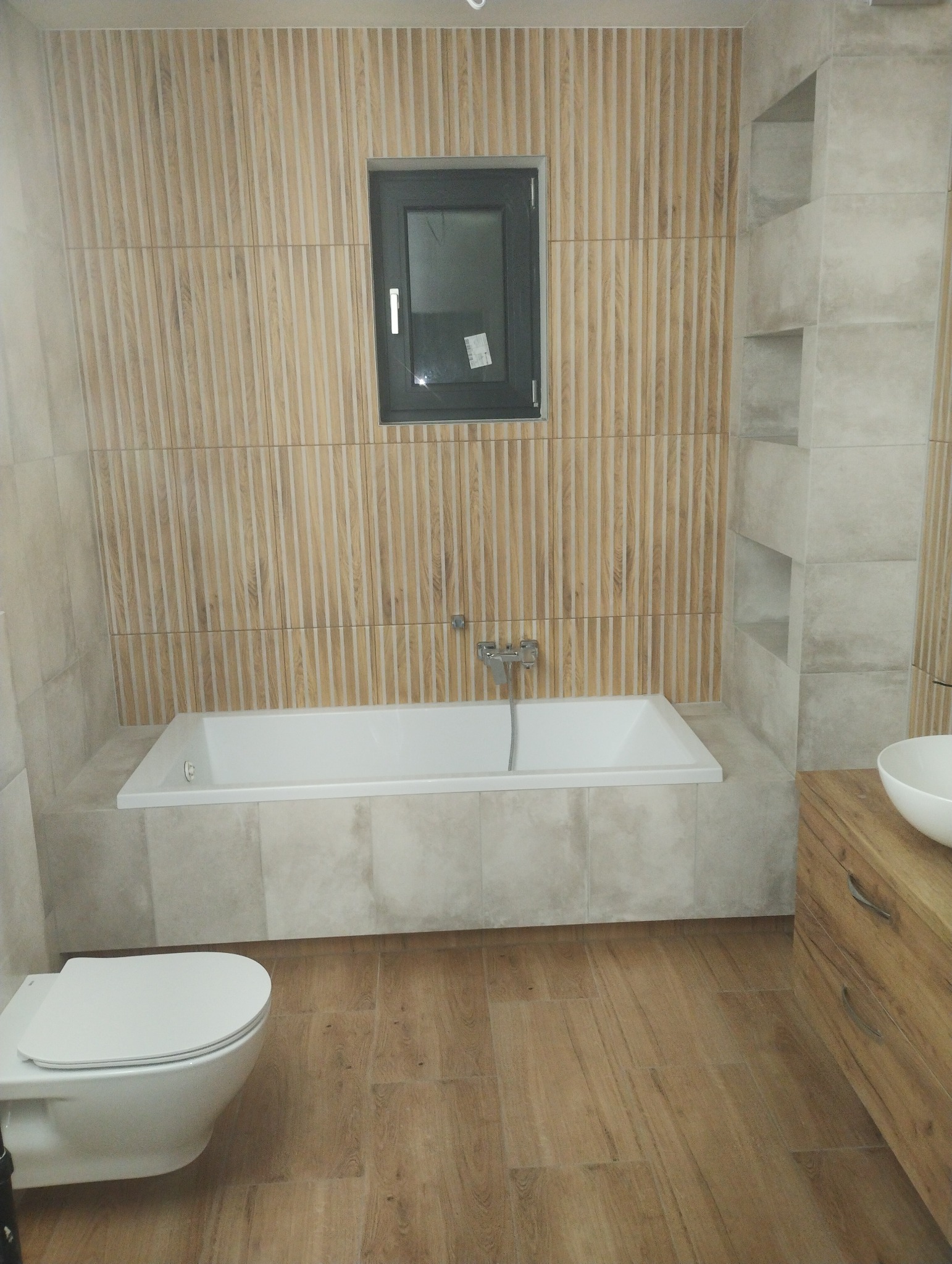 Nowoczesna łazienka z drewnopodobnymi płytkami na ścianie i podłodze, biała wanna pod oknem, wisząca toaleta i umywalka na drewnianej szafce.