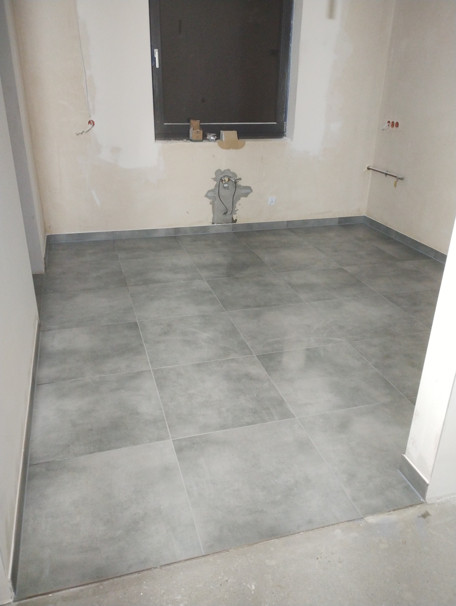 Szare płytki podłogowe ułożone w kwadraty w pustym pomieszczeniu z oknem i instalacjami na ścianach, widoczne niedokończone wykończenie ścian i podłogi.