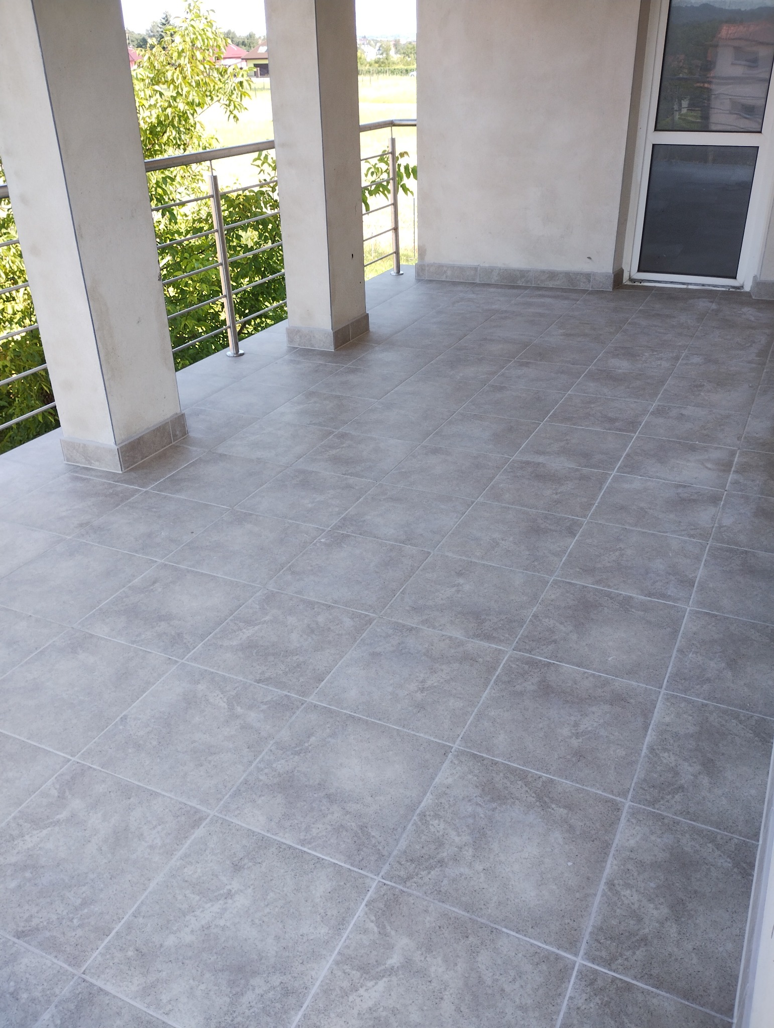 Szare płytki gresowe ułożone na balkonie z metalową balustradą i widokiem na zieleń, wykończenie z szarymi cokołami.