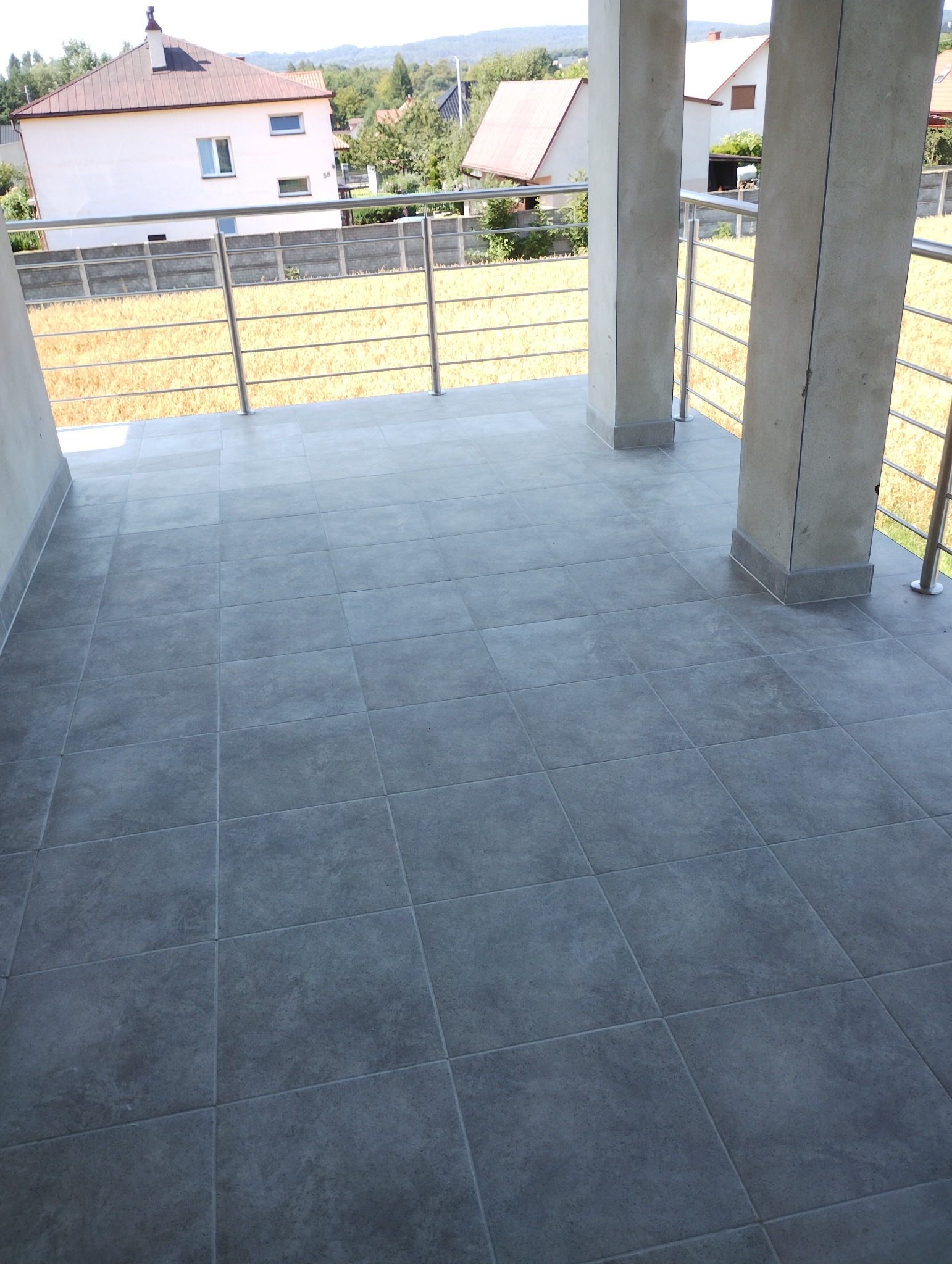 Szare płytki gresowe ułożone na balkonie z metalową balustradą i widokiem na domy w oddali.