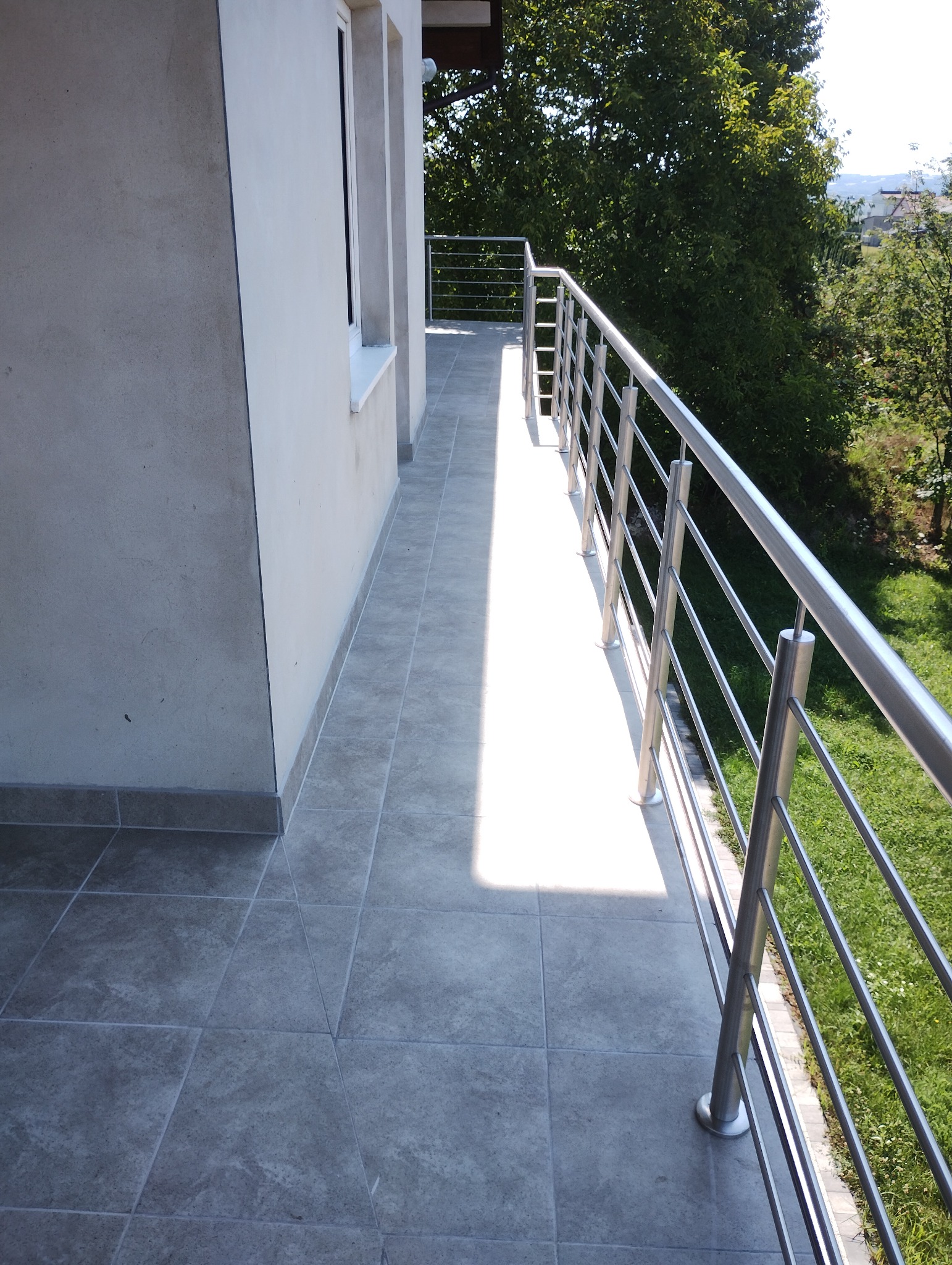 Wykończony balkon z szarymi płytkami i metalową balustradą, widok na zieleń w tle.