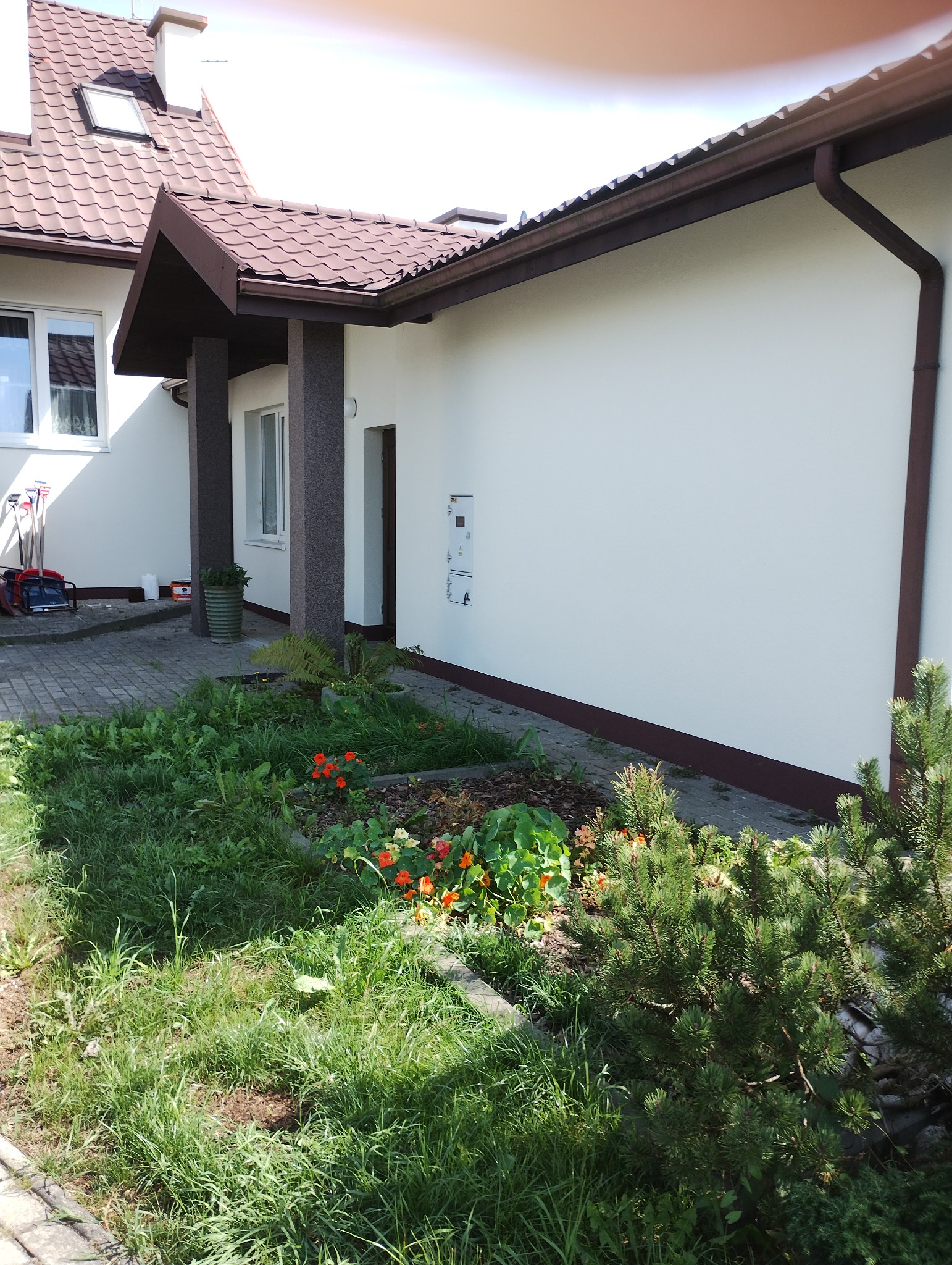 Jasna elewacja domu jednorodzinnego z brązowym dachem i elementami wykończeniowymi, widok z poziomu ogrodu z kwitnącymi nasturcjami.