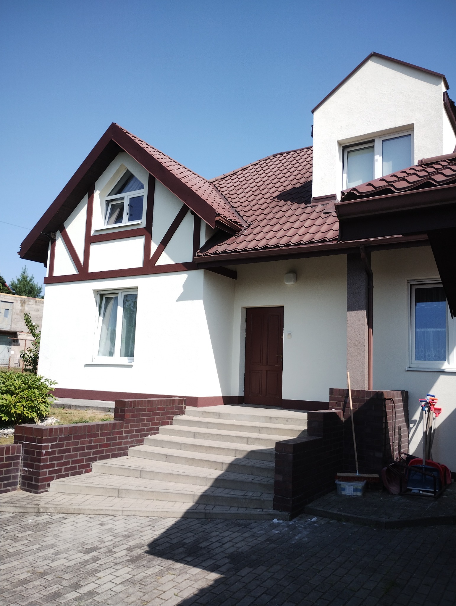 Elewacja domu jednorodzinnego w stylu muru pruskiego z brązowym dachem i schodami wejściowymi. Widoczne narzędzia sugerują prace wykończeniowe.
