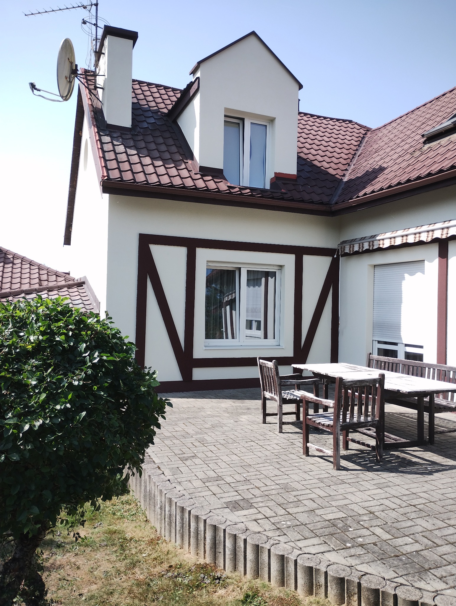 Elewacja domu z brązowymi elementami dekoracyjnymi, brązowy dach, okna z roletami. Na tarasie drewniany stół z krzesłami.