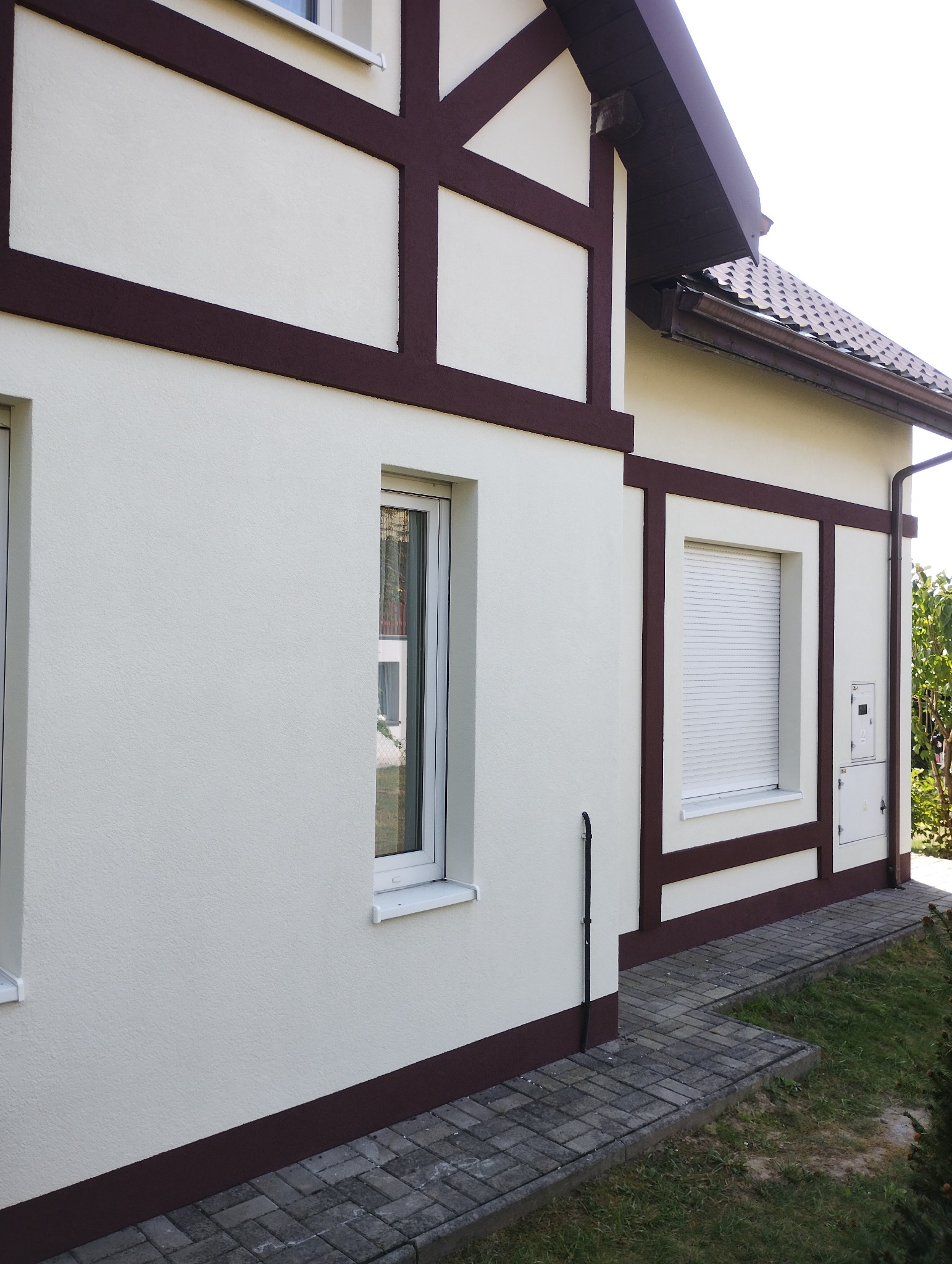 Elewacja domu z oknami i brązowymi elementami dekoracyjnymi, widok z boku, kostka brukowa przed budynkiem, jasne tynki.