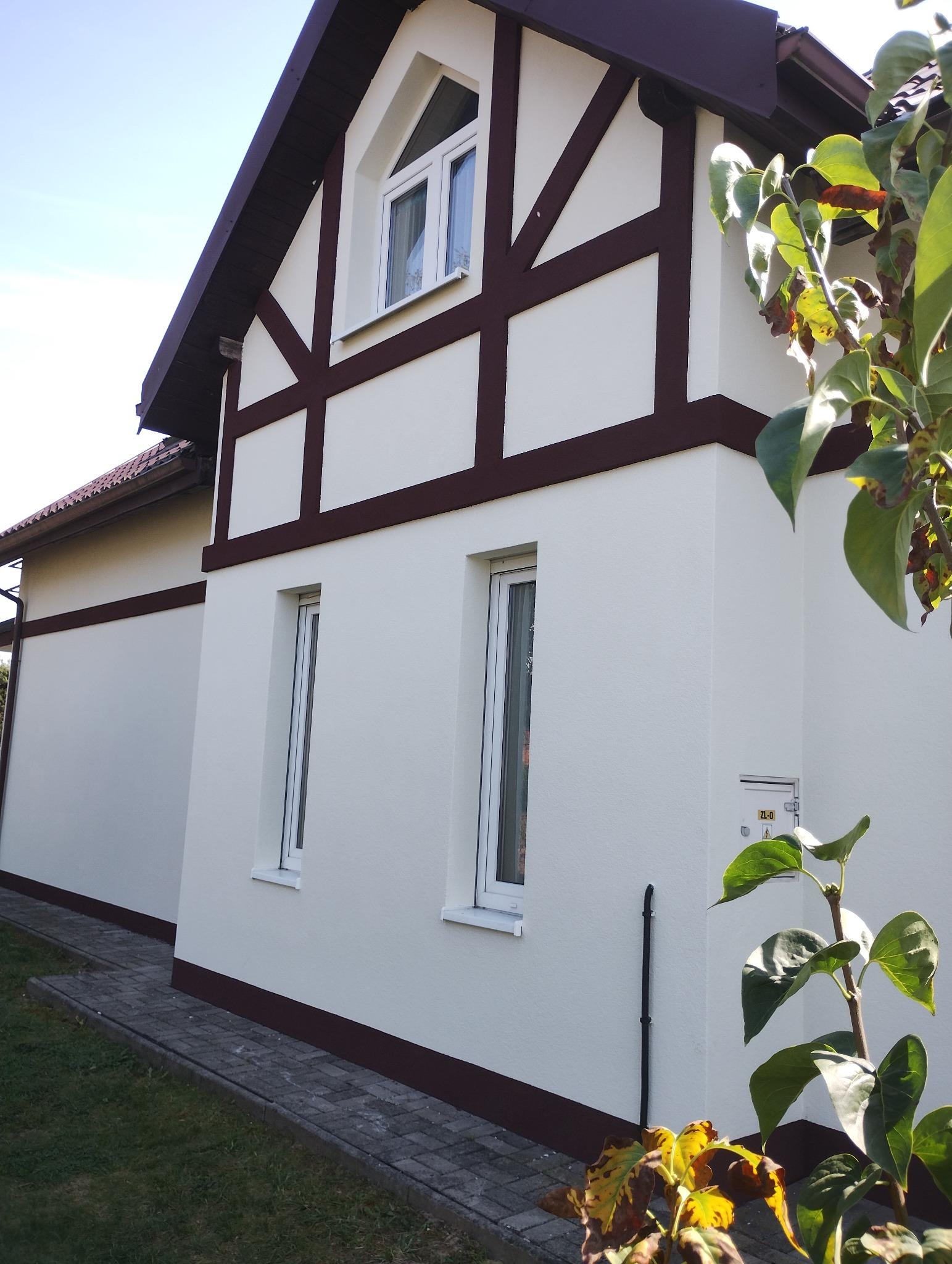 Odnowiona elewacja domu w stylu szachulcowym, z kremową ścianą i brązowymi belkami. Widoczne okna i fragment chodnika. Całość w słoneczny dzień.