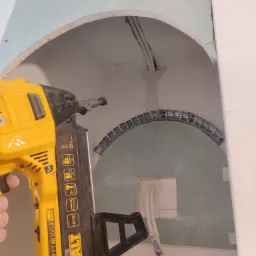 Ż&oacute;łta wkrętarka DeWalt przy mocowaniu płyty karton gips z wyciętym łukiem i widocznymi przewodami elektrycznymi.