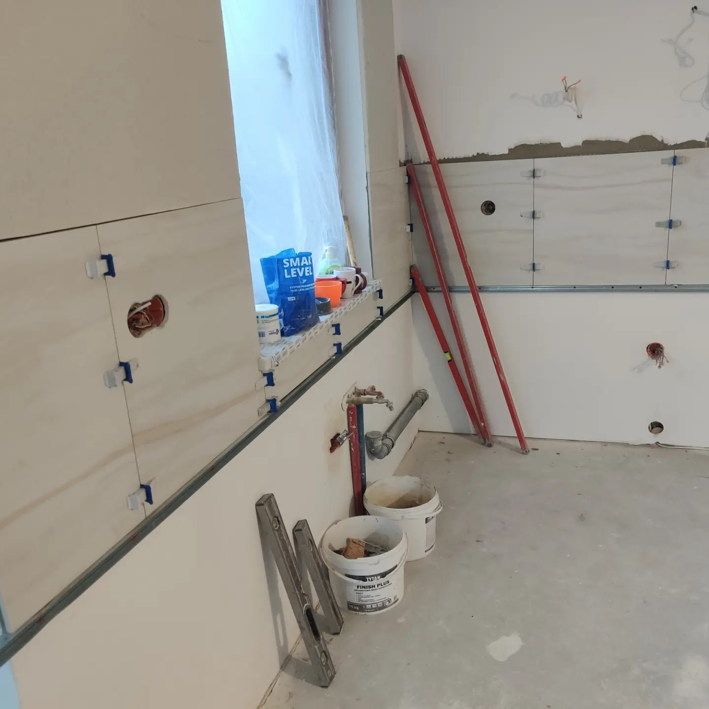 Montaż jasnych płytek ściennych w kuchni, widoczne poziomice, plastikowe krzyżyki dystansowe, puszki elektryczne i wiadro z zaprawą klejową