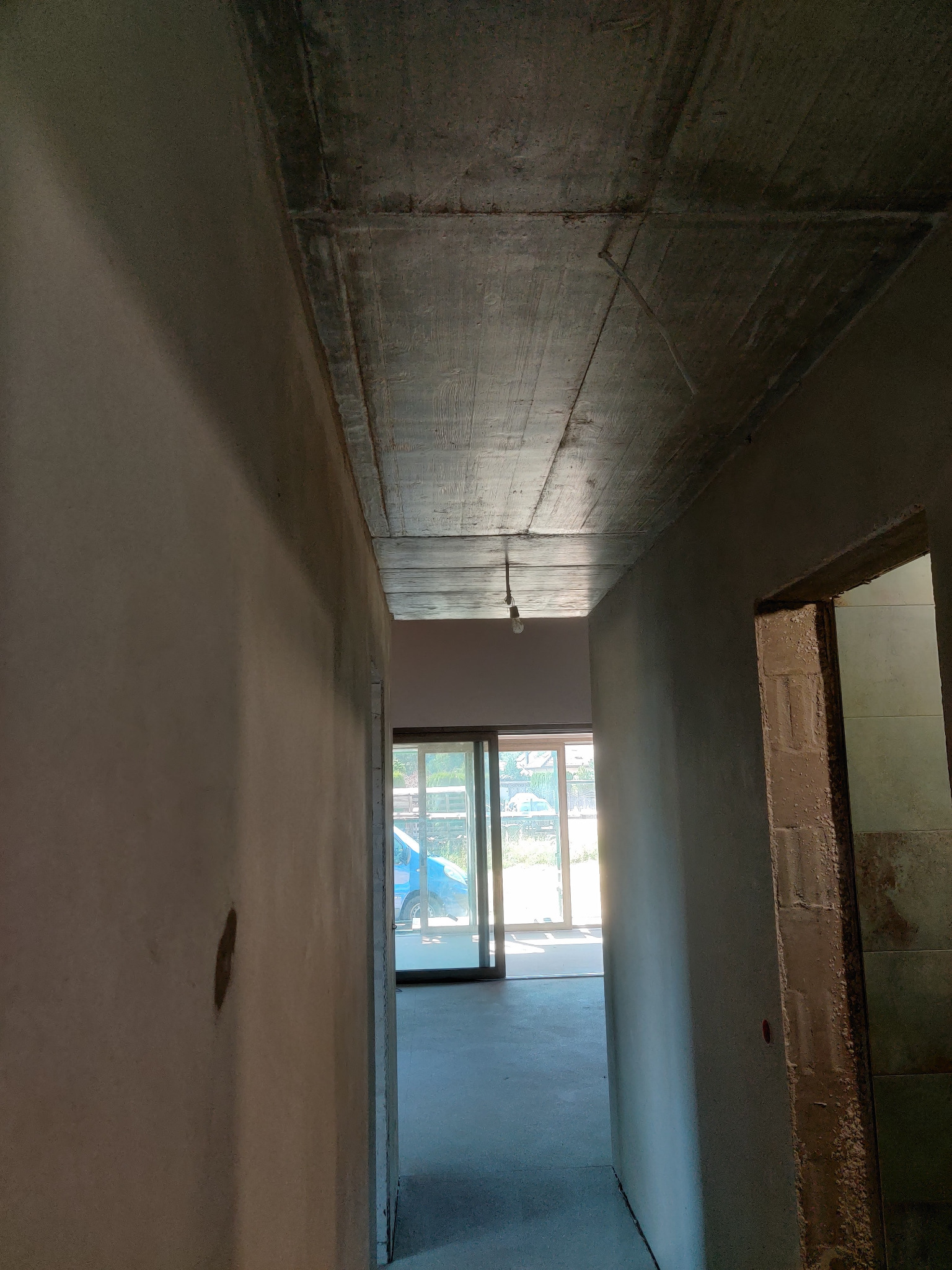 Wnętrze w trakcie prac wykończeniowych: surowy betonowy strop, otynkowane ściany z widocznymi niedoskonałościami, surowa ościeżnica, w tle przesuwne drzwi balkonowe i słabe oświetlenie żarówką...